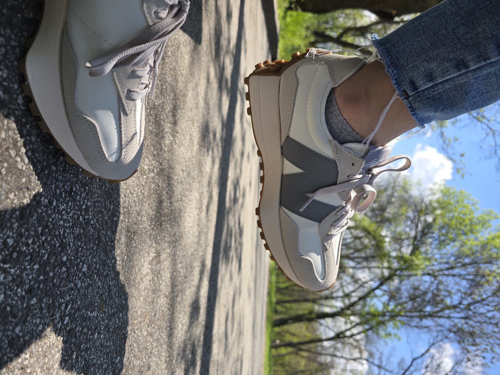 The image captures a close-up, low-angle view of a person's feet and lower legs, rotated 90 degrees clockwise. The person is wearing light-colored sneakers, blue distressed jeans with frayed hems, and gray ankle socks. The sneaker prominently displayed on the right features a white and light grey upper with a distinct dark grey "N" logo on the side, typical of New Balance footwear. It has light grey laces and a visible brown, ridged sole extending up the heel. The left shoe is partially visible, showing similar light colors and texture.
The setting appears to be outdoors on a sunny day. Long, distinct shadows are cast on the paved ground, which is a mix of dark and lighter asphalt or concrete, suggesting either morning or late afternoon. In the background, lush green trees with emerging leaves are visible against a bright blue sky with scattered white clouds. The overall scene suggests a leisurely walk or stand in a park, on a sidewalk, or a quiet street. The activity is implied as standing or walking. There is no other discernible text beyond the "N" logo on the shoe.