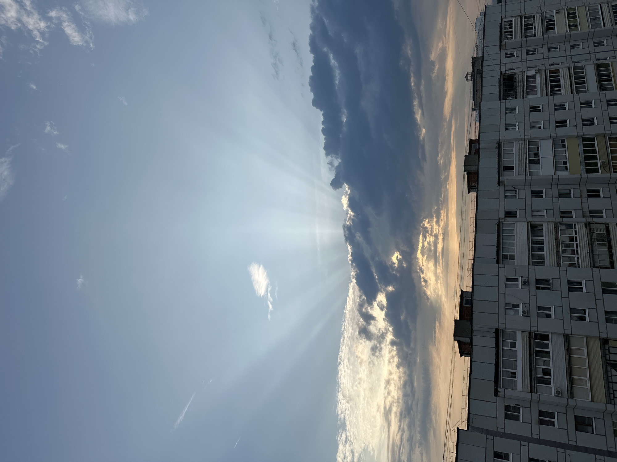 This is a photograph of a residential apartment building in the city of Chaikovsky, Russia. The building appears to be made of concrete and is several stories high, with numerous windows visible. The sky above is a mix of blue and grey clouds, with sunbeams radiating from behind the clouds. The lighting suggests it is either sunrise or sunset. The photograph is taken from a slightly elevated perspective, looking up at the building and the sky.