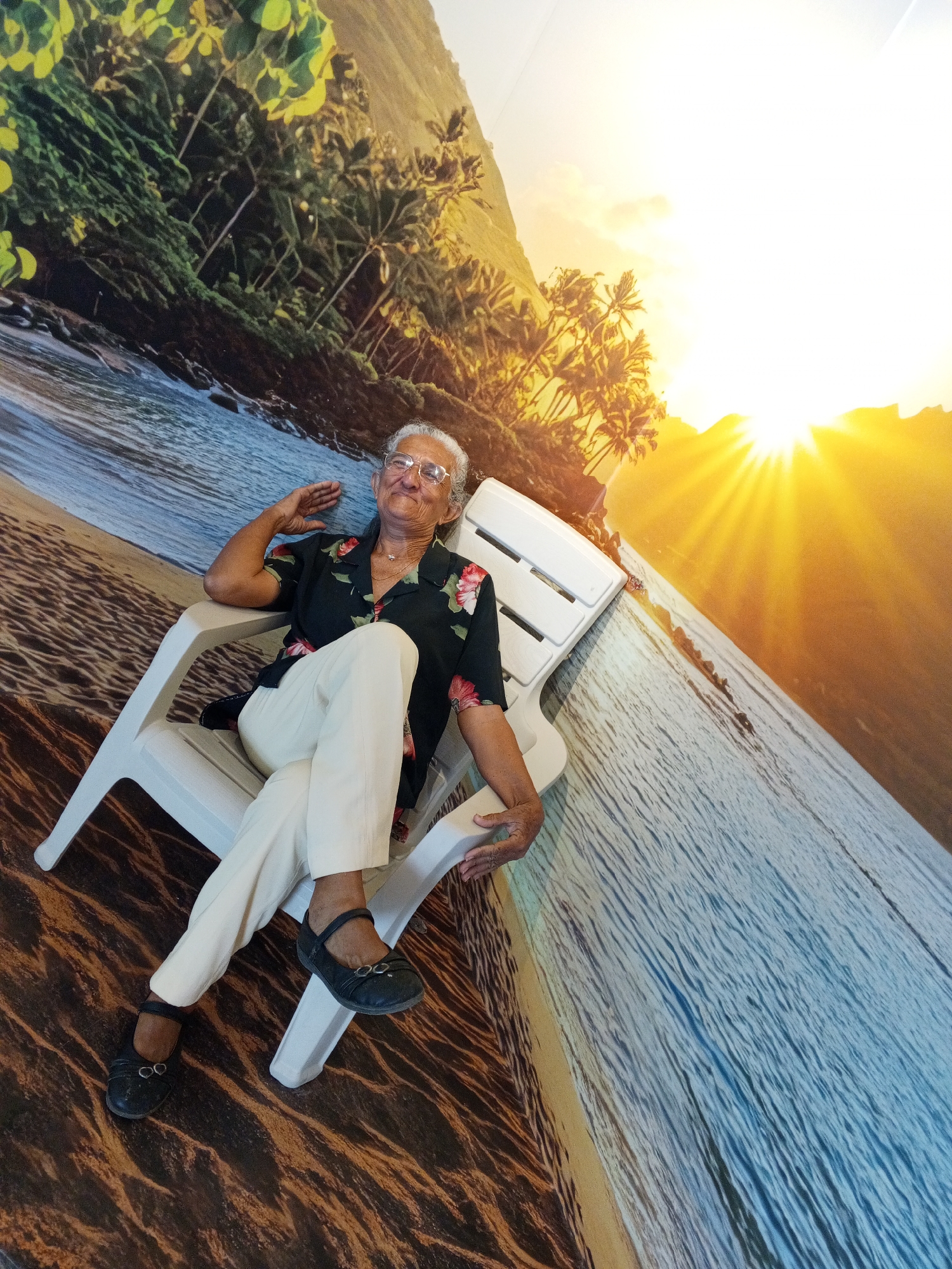 An elderly woman with gray hair and glasses sits in a white plastic lounge chair in front of a mural of a tropical beach scene. The mural depicts palm trees, a mountain, a sandy beach, and ocean water under a bright, setting sun. The woman is wearing a dark floral shirt with pink and red flowers, cream-colored pants, and black flat shoes. She is leaning back in the chair, with her left arm resting on the armrest and her right hand raised near her head, as if shielding her eyes from the sun or posing. Her legs are crossed. The chair is positioned on the sandy part of the mural.