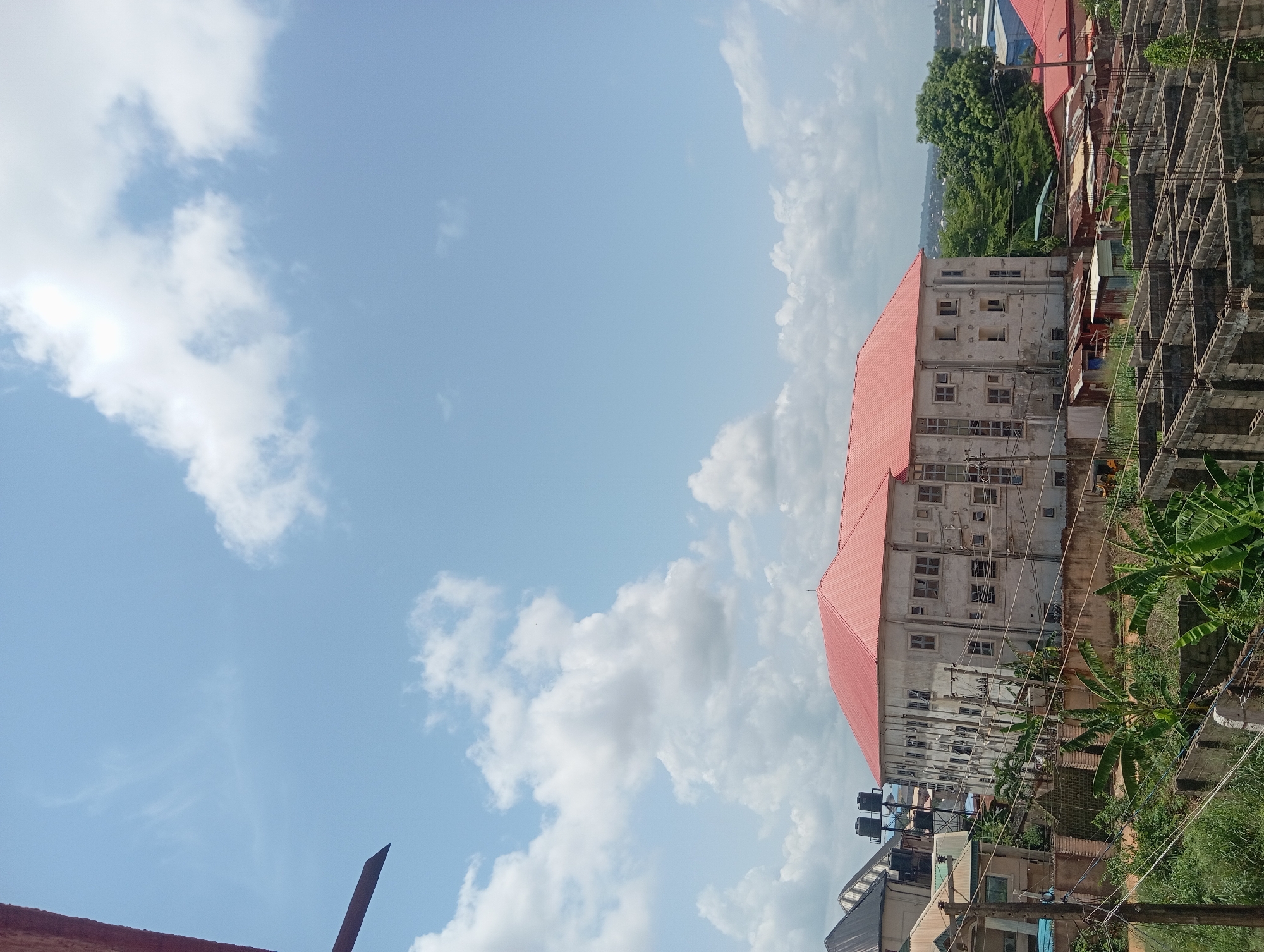 The image, oriented sideways, depicts a partially cloudy sky above a built environment in Nkpor, Nigeria. A large, multi-story building with a red roof dominates the midground. The building appears worn, with numerous windows and signs of weathering. Adjacent to the building is an unfinished concrete structure with exposed rebar. Dense green vegetation, including banana trees and other plants, is visible in the foreground and around the buildings. Overhead wires are present. In the background, there are hints of other buildings and greenery. The sky is mostly blue with scattered white clouds.