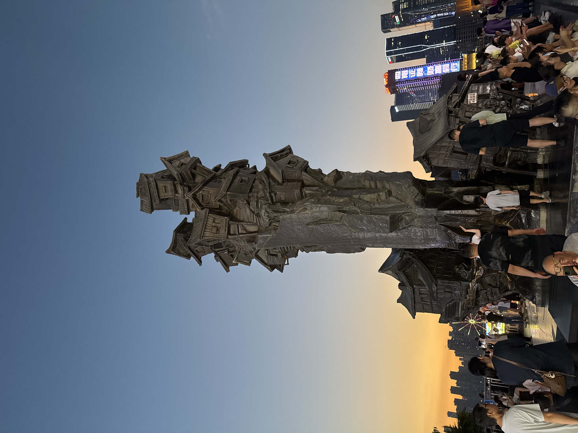 The image captures a bronze sculpture resembling a cluster of houses precariously stacked upon a rocky outcrop, set against the backdrop of a city skyline at dusk. The sculpture is the central focus, with its detailed architectural elements and weathered texture. A clear sky transitions from a light blue overhead to a warm orange near the horizon, indicating the setting sun. In the foreground and midground, a diverse group of people are present, some standing, some sitting, and many engaged with their mobile phones. They are gathered around the sculpture, suggesting it's a public landmark or attraction. Behind the sculpture, modern high-rise buildings with illuminated windows and signs are visible, contrasting with the traditional-style houses depicted in the artwork. A few palm trees are also present to the left of the frame. The scene appears to be a waterfront area, possibly a promenade or plaza, given the open space and the presence of multiple individuals. The overall impression is one of a vibrant urban public space at the end of the day, with a significant artistic centerpiece drawing people's attention.