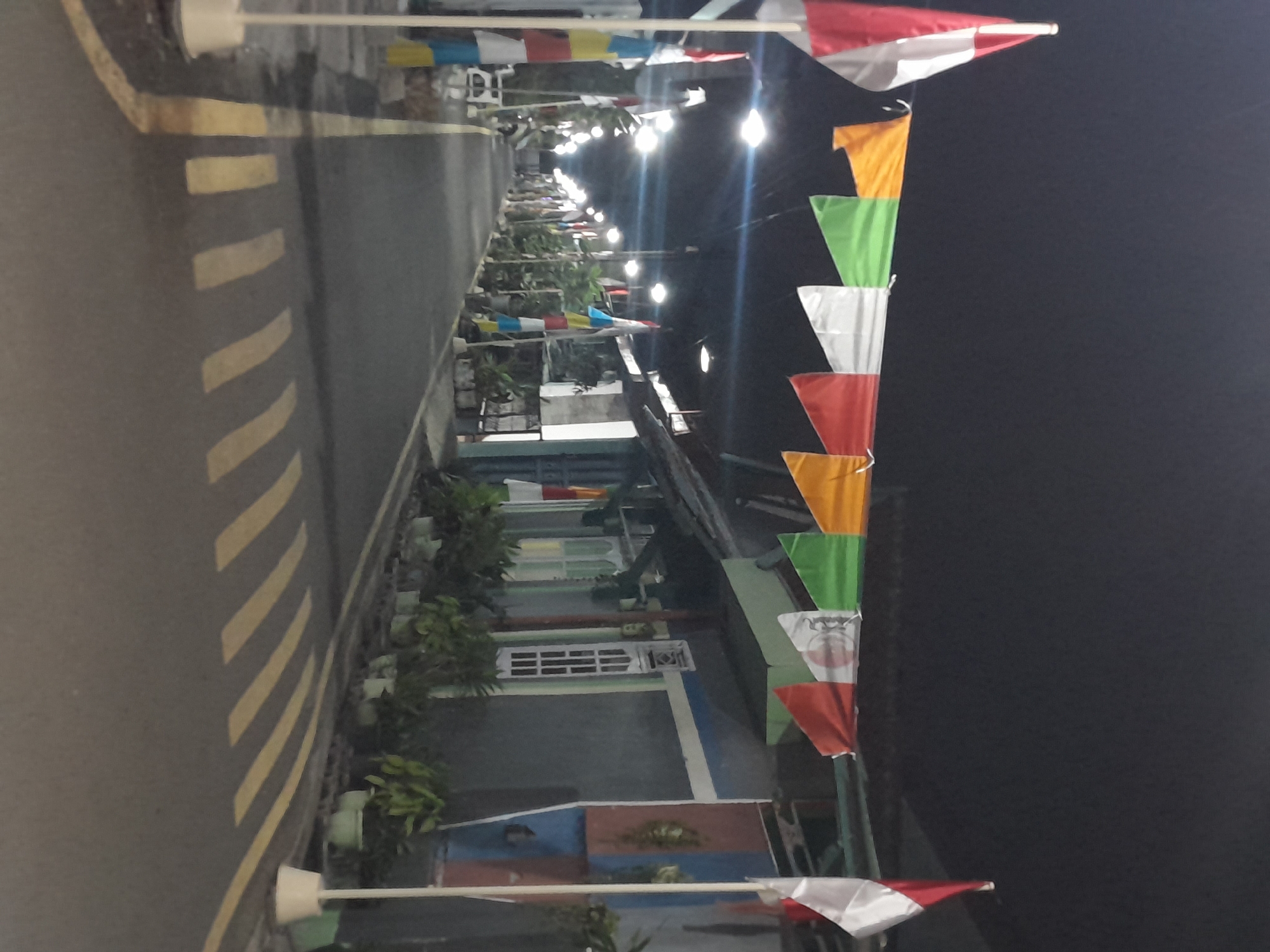 The image depicts a street in Mejasem Timur, Indonesia, at night. The street is lined with houses on both sides, and decorative flags are strung across the road at intervals. These flags are triangular and appear to be in various colors, including red, white, orange, and green. Some flags also feature patterns or symbols.

Along the sides of the street, there are streetlights illuminating the scene. The road itself is paved and has yellow markings, indicating lanes or pedestrian crossings. Potted plants are visible in front of the houses, adding some greenery to the environment. The houses have a simple architectural style.

On the left side of the image, there are poles with more flags attached vertically. These flags also appear to be red and white.

There are no people visible in the image. The overall impression is that of a festive or celebratory atmosphere, suggested by the colorful flags decorating the street. The lighting indicates it is nighttime, possibly after dark.