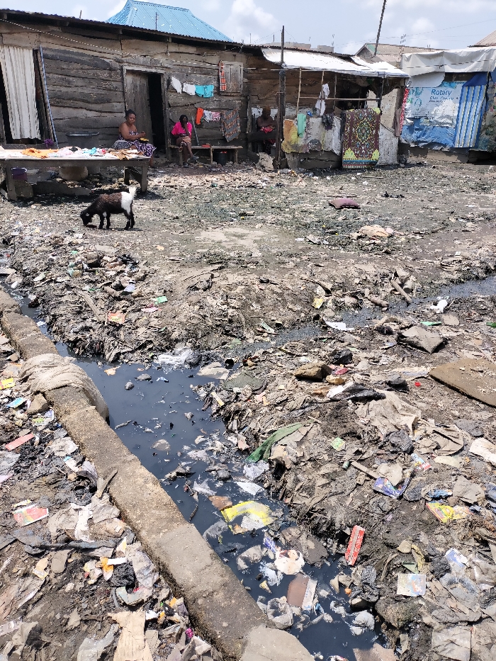 This image depicts a densely packed informal settlement or slum in Oworonshoki, Nigeria, characterized by significant environmental degradation. In the foreground, an open drainage channel runs horizontally, filled with dark, stagnant water heavily contaminated with various solid waste items, including plastics, paper, and unidentified debris. A concrete or wooden beam, partially wrapped with fabric, delineates the left bank of this channel. The ground immediately surrounding the channel and extending into the midground is an uneven expanse of compacted dirt and mud, heavily littered with trash.
In the mid-left, a small, black and white goat actively forages on the refuse-strewn ground. The background features a row of closely built, single-story structures. The most prominent building on the left is constructed from rough, horizontally stacked wooden planks, topped with a light blue corrugated metal roof. Two open doorways are visible; the left one partially obscured by a white curtain. An adult female is seated on a low wooden bench or platform outside the central door, facing the viewer, wearing patterned clothing. A white bowl is placed near her. Further to her right, another individual, possibly female, is seated on a similar bench, clad in bright pink. A third, less distinct person is visible further right. Clothes and patterned fabrics are hung to dry on lines attached to these structures and an adjacent makeshift stall.
To the right of the wooden structure, additional makeshift dwellings or stalls are constructed from a variety of materials, including wood, corrugated metal, and blue tarpaulins. One blue tarpaulin prominently displays a "Rotary Club" logo and text. The sky overhead is partly cloudy, indicating daytime conditions.