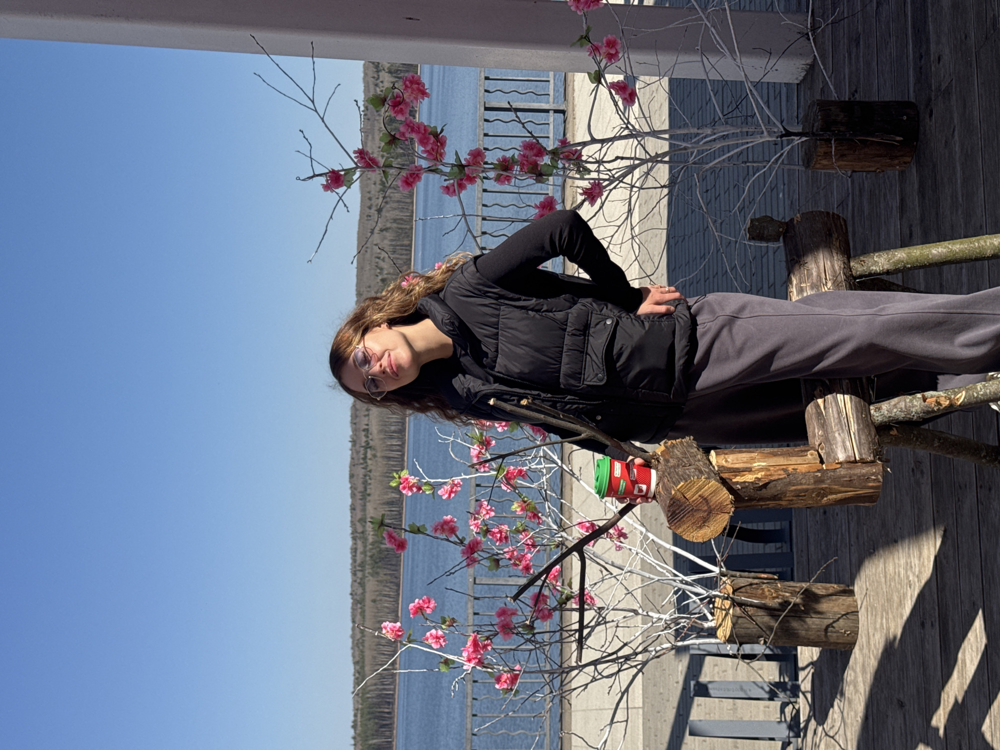 A woman with long brown hair and glasses stands on a wooden deck next to a faux flowering tree, holding a paper cup. She wears a black puffer vest over a black long-sleeve shirt and gray sweatpants. Behind her is a body of water and a wooded shoreline under a blue sky. The tree is decorated with pink flowers and is placed in a wooden base. The deck has gray metal railings. The location is identified as the city of Chaikovsky, Russia, based on visual features.