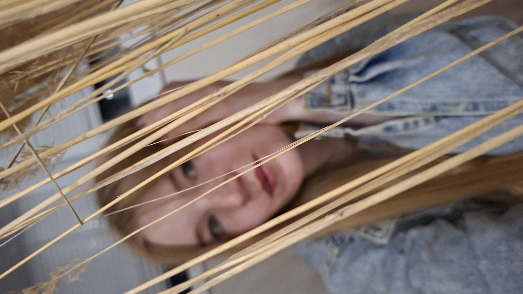 The image shows a medium shot of a young woman with long, blonde hair, looking towards the camera through dry, light-brown reeds. She is wearing a light-wash denim jacket. Her left hand is near her face, touching her cheek and forehead. The background is blurred, showing a light-colored wall and a dark rectangle, possibly a window or door, with a small, bright circular reflection within it. The scene appears to be indoors, in an urban environment. There is no specific indication of the city of Chaikovsky, Russia, within the image.