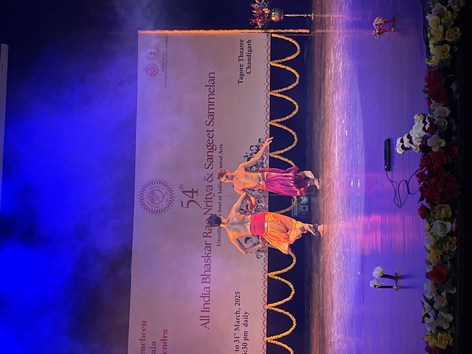 The image depicts a performance of the 54th All India Bhaskar Rao Nritya & Sangeet Sammelan at the Tagore Theatre in Chandigarh, India (latitude: 30.733, longitude: 76.79). Two male dancers wearing traditional Indian clothing are performing a dance routine on a stage illuminated by purple and pink lights. The backdrop features the event's name and logo, as well as a date and time: "1st to 31st March, 2025, 6:30 pm daily." The stage is adorned with yellow garlands. The stage is also decorated with floral arrangements and a black microphone on the floor. The event is supported by the Ministry of Culture, Government of India.