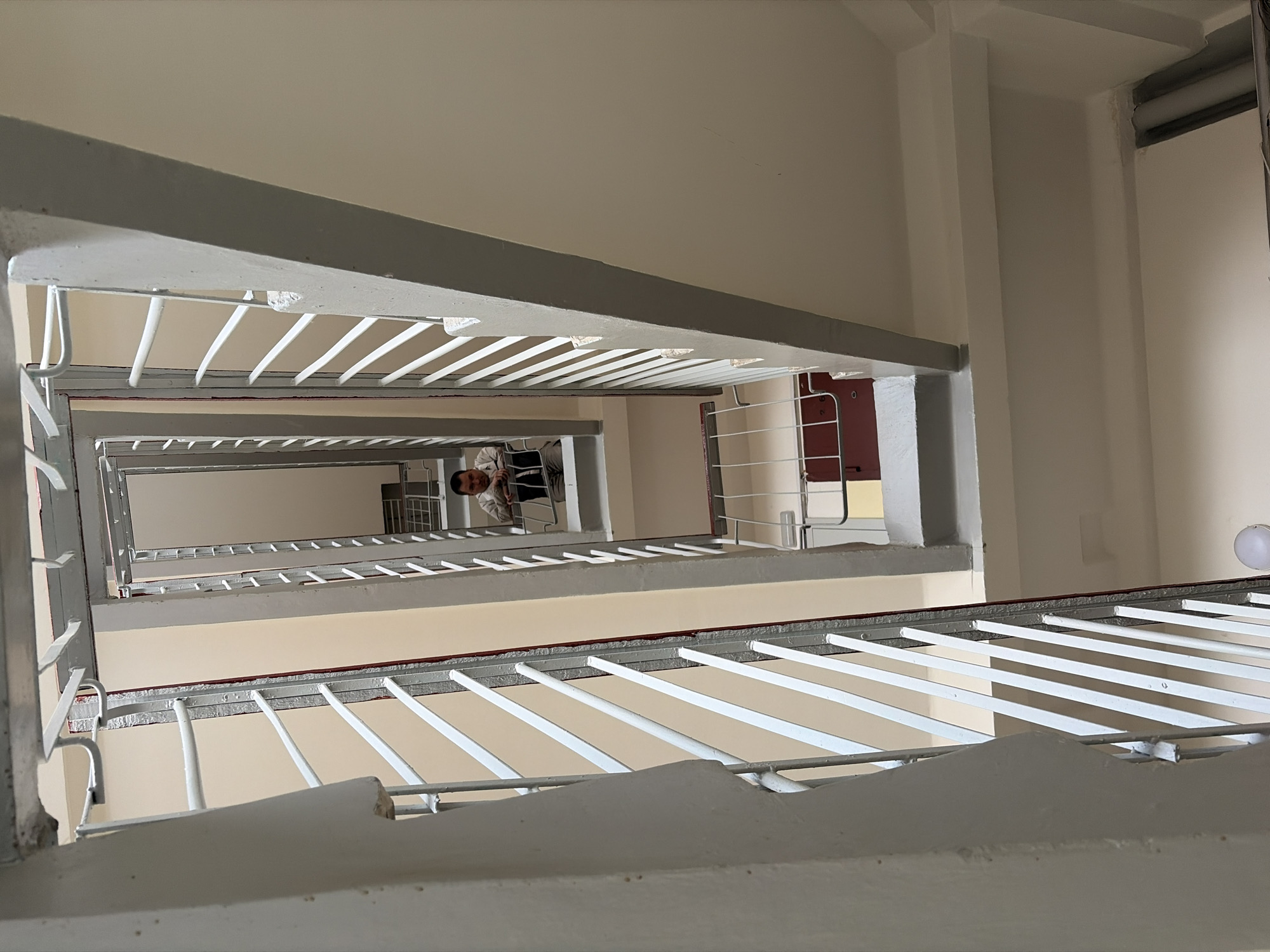 The image depicts an interior view looking up a stairwell in a building. The stairwell walls are primarily beige and white. The stairs are concrete with white metal railings. A man is visible on an upper floor. The presence of the staircase, paint colors, and railings are indicative of the location in Izhevsk, Russia.