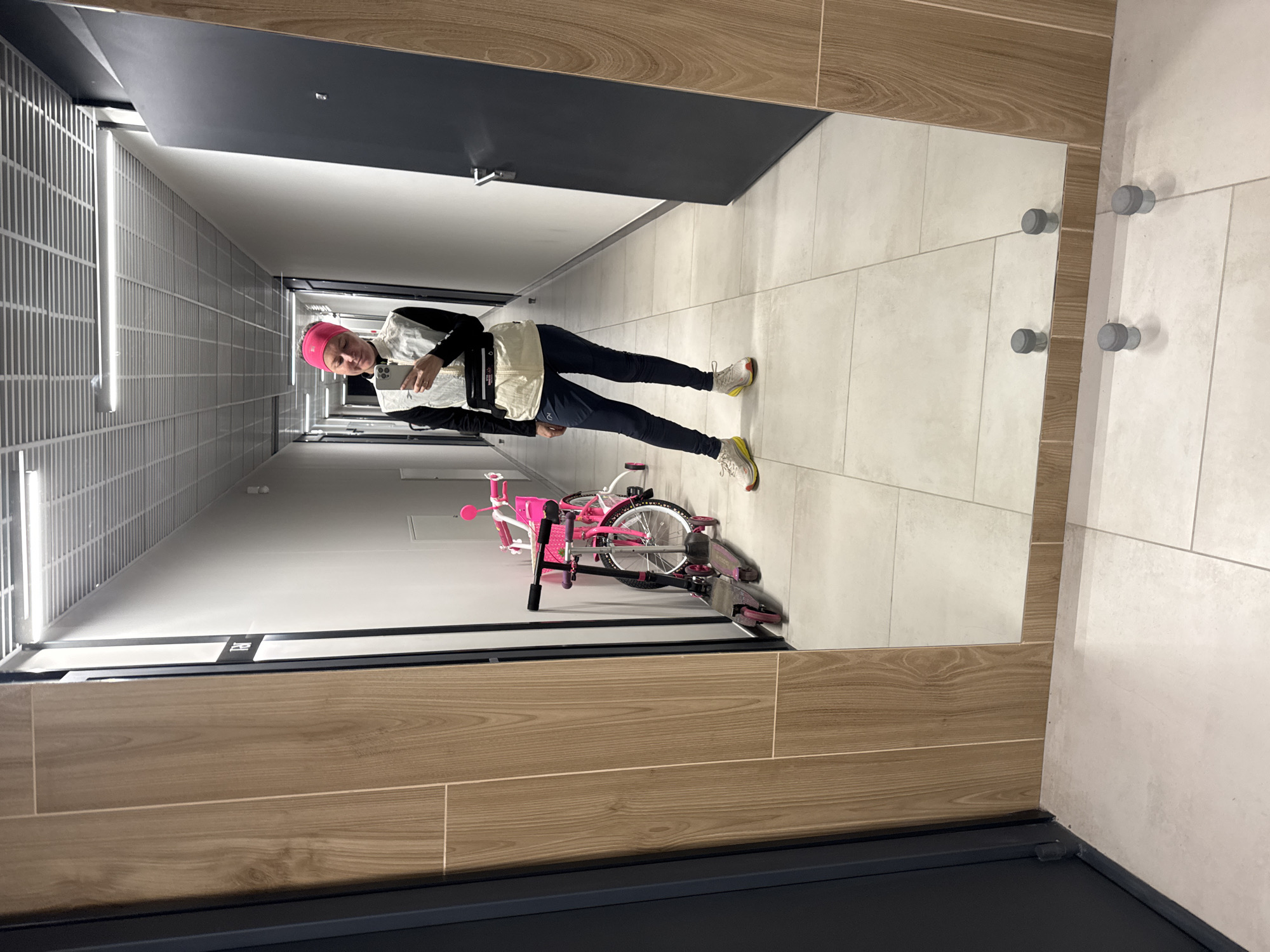 A person with a pink head covering is standing in a hallway holding a mobile phone. The hallway has a bike leaning against a wall, with doors on one side and a tiled wall on the other. The image appears to be a reflection in a mirror. The location is indeterminate, and the details of the scene do not provide evidence to confirm Verkhnii Liuk, Russia.