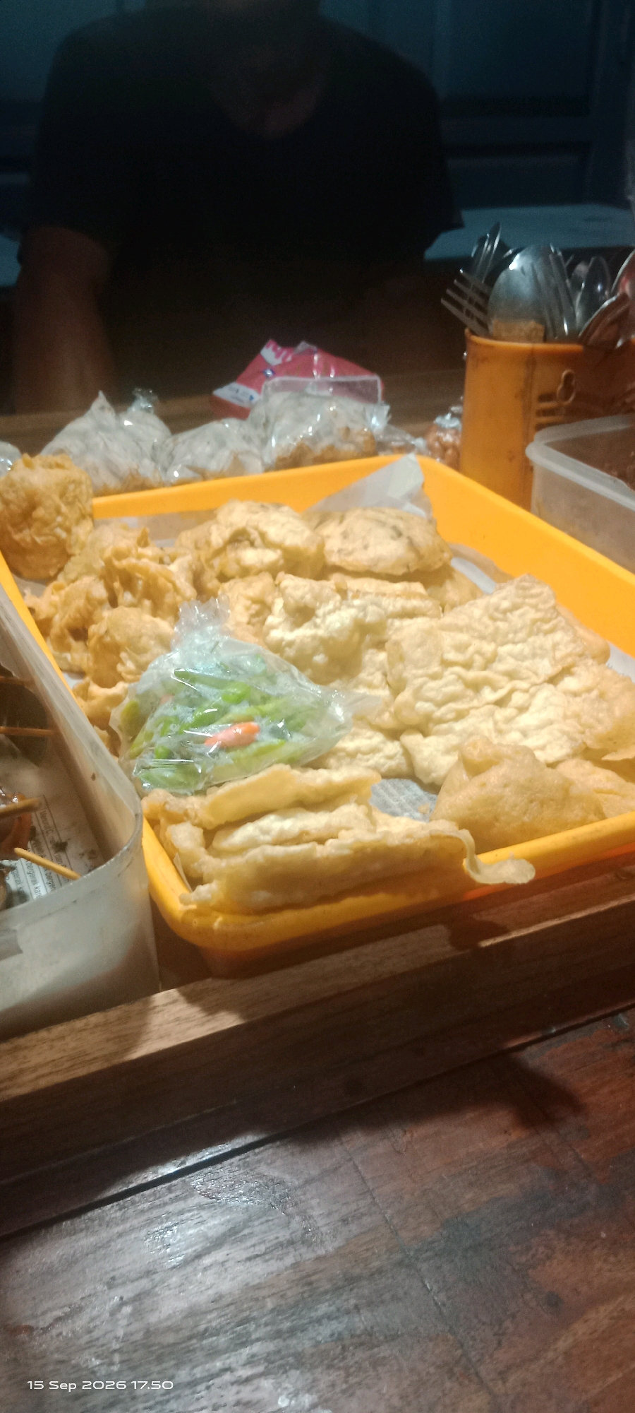 A tray of fried food is on a wooden table. The tray is orange and filled with various fried items, including what appears to be fried tofu or tempeh, and some round, golden-brown pieces that could be fried meatballs or fish balls. In the center of the tray, there is a clear plastic bag containing green chilies and what looks like a red chili. Behind the tray, there are also some clear plastic bags containing more food items. To the left of the tray, there is a white plastic container holding skewers of food. In the background, out of focus, a person wearing a dark t-shirt is visible. To the right of the tray, a yellow container holds cutlery. The image was taken in Merbung, Indonesia.