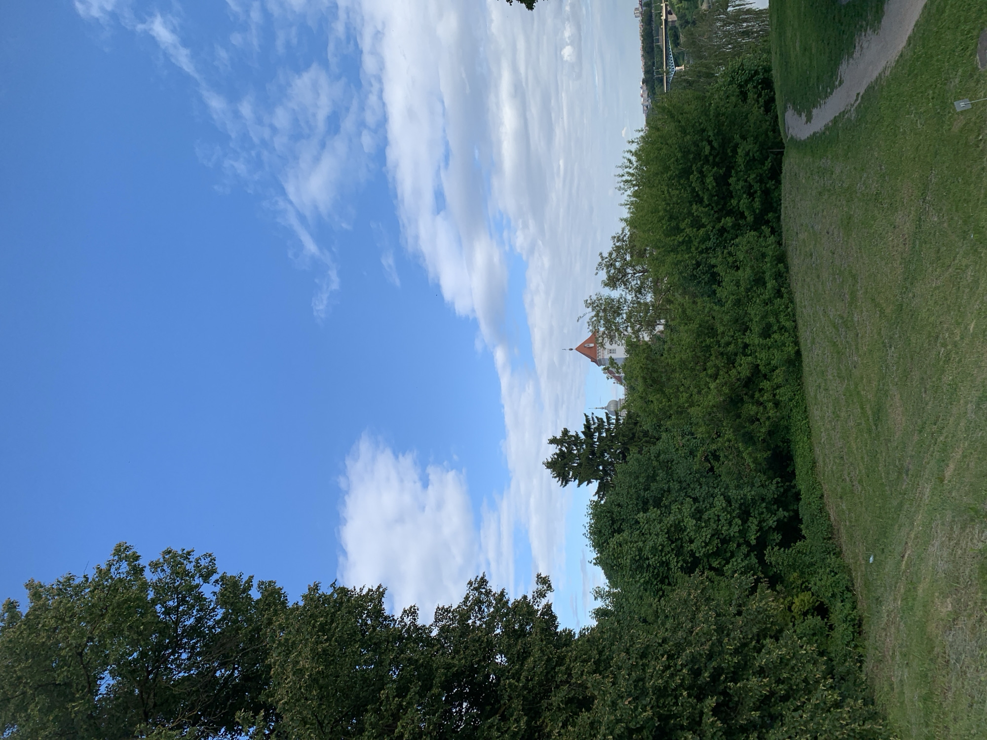 The image captures a view in Hrodna, Belarus, featuring a red-roofed building partially visible amidst dense green trees. The scene includes a patch of green grass, a winding path, and a bridge spanning a body of water in the background. The sky is a blend of blue and white clouds, contributing to the overall naturalistic setting of the scene.