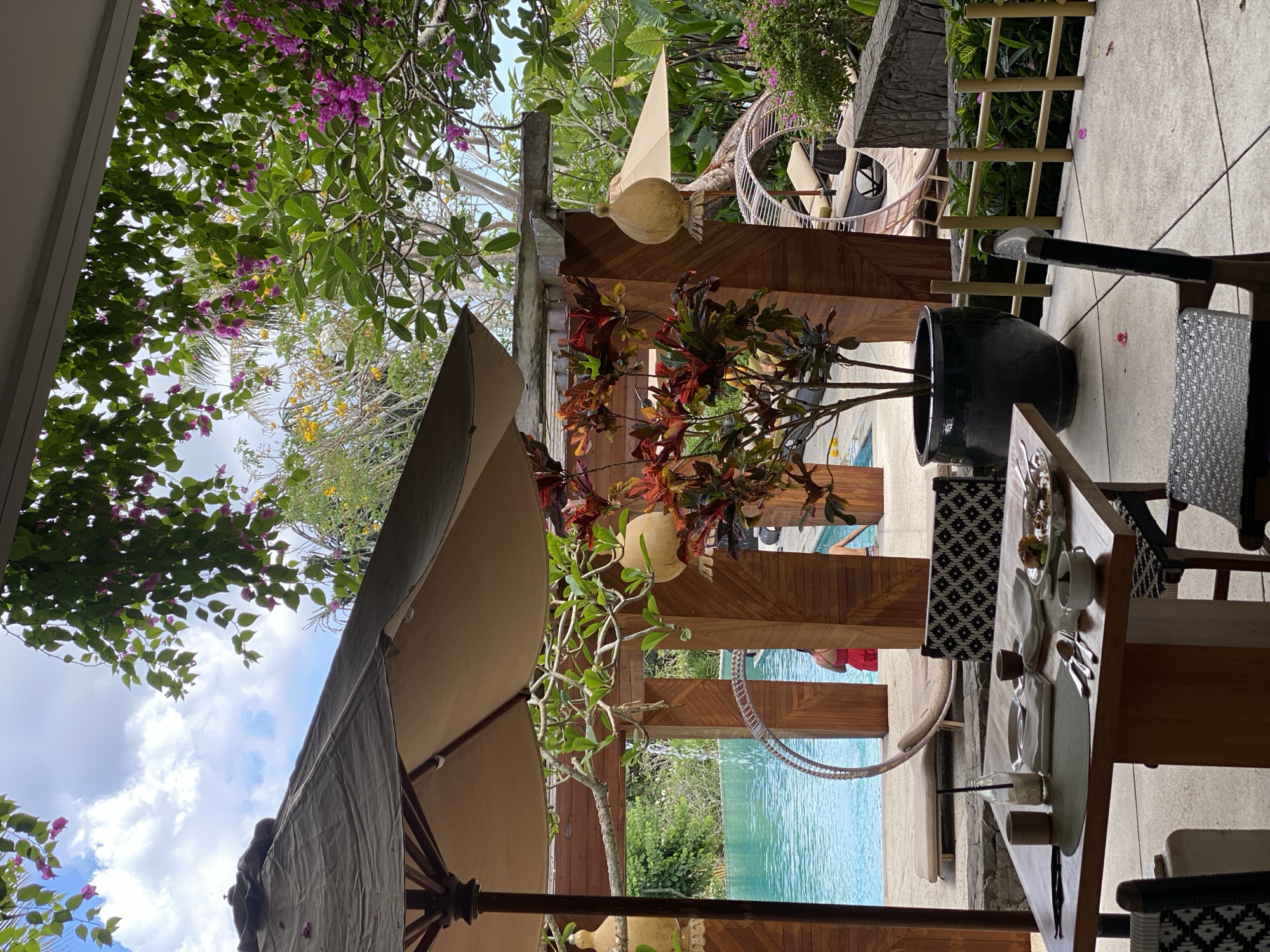 The image depicts an outdoor dining area with a swimming pool in the background. A wooden table is set for a meal with plates, cutlery, and cups. Two chairs are visible at the table, one in the foreground and another partially visible. Behind the table, a large potted plant with red and green leaves stands. In the background, a swimming pool with clear blue water is bordered by lounge chairs and a wooden structure. Lush green foliage and pink flowers are abundant throughout the scene. The location is identified as Kuta Selatan, Indonesia.