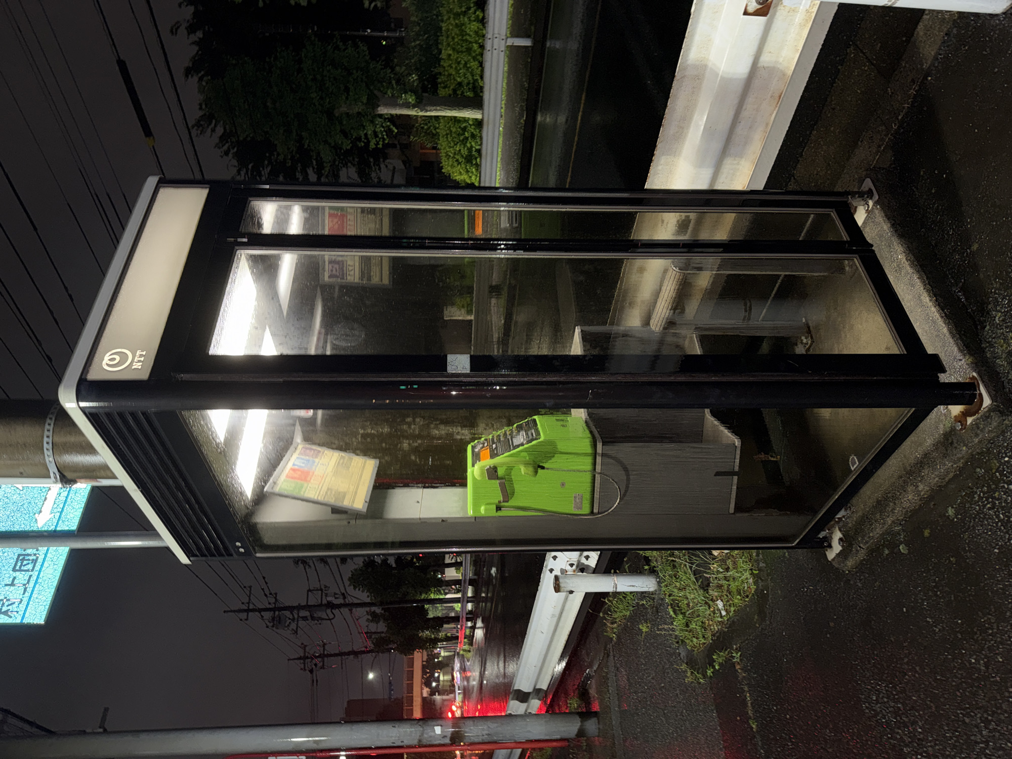 The image depicts a public telephone booth on a wet street in Yachiyo, Japan. The booth is made of black metal and glass, with a light-up sign at the top displaying the "NTT" logo. Inside, a bright green payphone is mounted on the wall, with a coiled cord hanging down. The street is wet and reflective, indicating recent rain. Traffic lights and streetlights are visible in the background, along with trees and other street furniture. The scene appears to be at night, with the phone booth being the primary source of light. The image is taken from a slightly low angle, looking up at the phone booth.