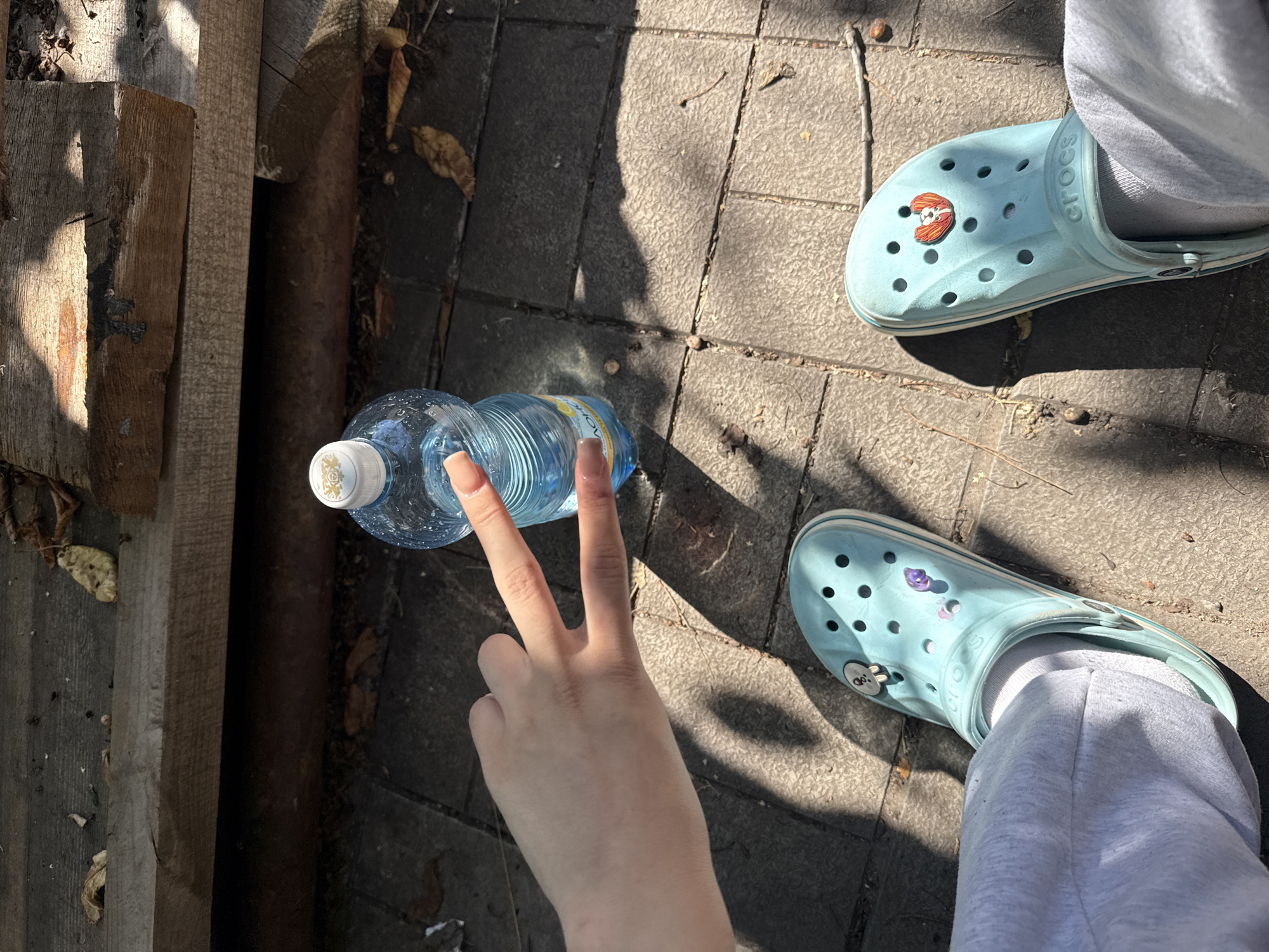 A person's legs and hands are visible, wearing light blue Crocs with charms and gray sweatpants. The person is holding a partially filled plastic water bottle, with their hand extended in a peace sign gesture. The scene is outdoors, with a ground made of gray interlocking pavers. Sunlight casts shadows, suggesting it is daytime with clear weather. To the left of the frame, there are pieces of unfinished wood and dried leaves scattered on the ground, hinting at a natural or perhaps a slightly neglected outdoor area. The shadows and the dry leaves suggest it might be autumn. The overall mood appears casual and relaxed. The word "CROCS" is visible on one of the shoes.