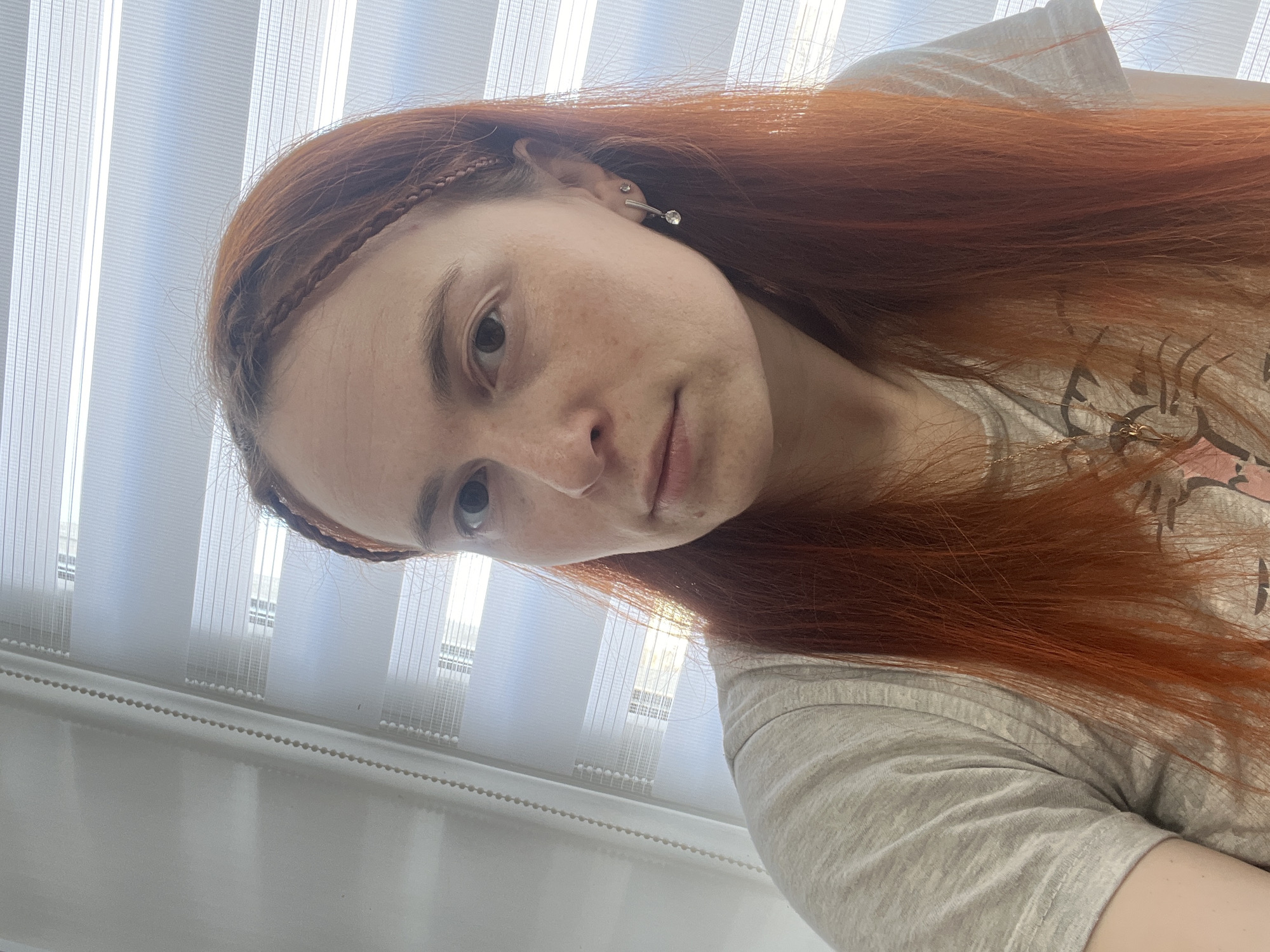 A portrait of a young person with long, reddish-brown hair, featuring small braids near the forehead. The person has fair skin, freckles, and visible earrings. They are wearing a light grey t-shirt. The background consists of vertical blinds. While the image's origin is stated as Chaikovsky, Russia, the photo itself provides no locational information beyond what is visible in the image.