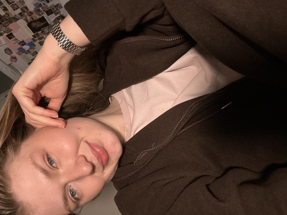 A female individual with light brown hair and blue eyes is depicted in a close-up, chest-up indoor portrait. She smiles directly at the camera, with her left hand positioned against her left cheek and ear area. A silver-toned metal link watch is visible on her left wrist. She wears a dark brown zippered hooded top, partially unzipped, over a light pink crew-neck t-shirt. The garment features a drawstring with a metal aglet visible on the right side. The background consists of a wall covered with multiple small square photographs and written notes. A white rectangular object is partially visible in the upper left background, and a red square object is present in the upper center-left. This scene is situated within an indoor residential environment in Kazan, Russia.