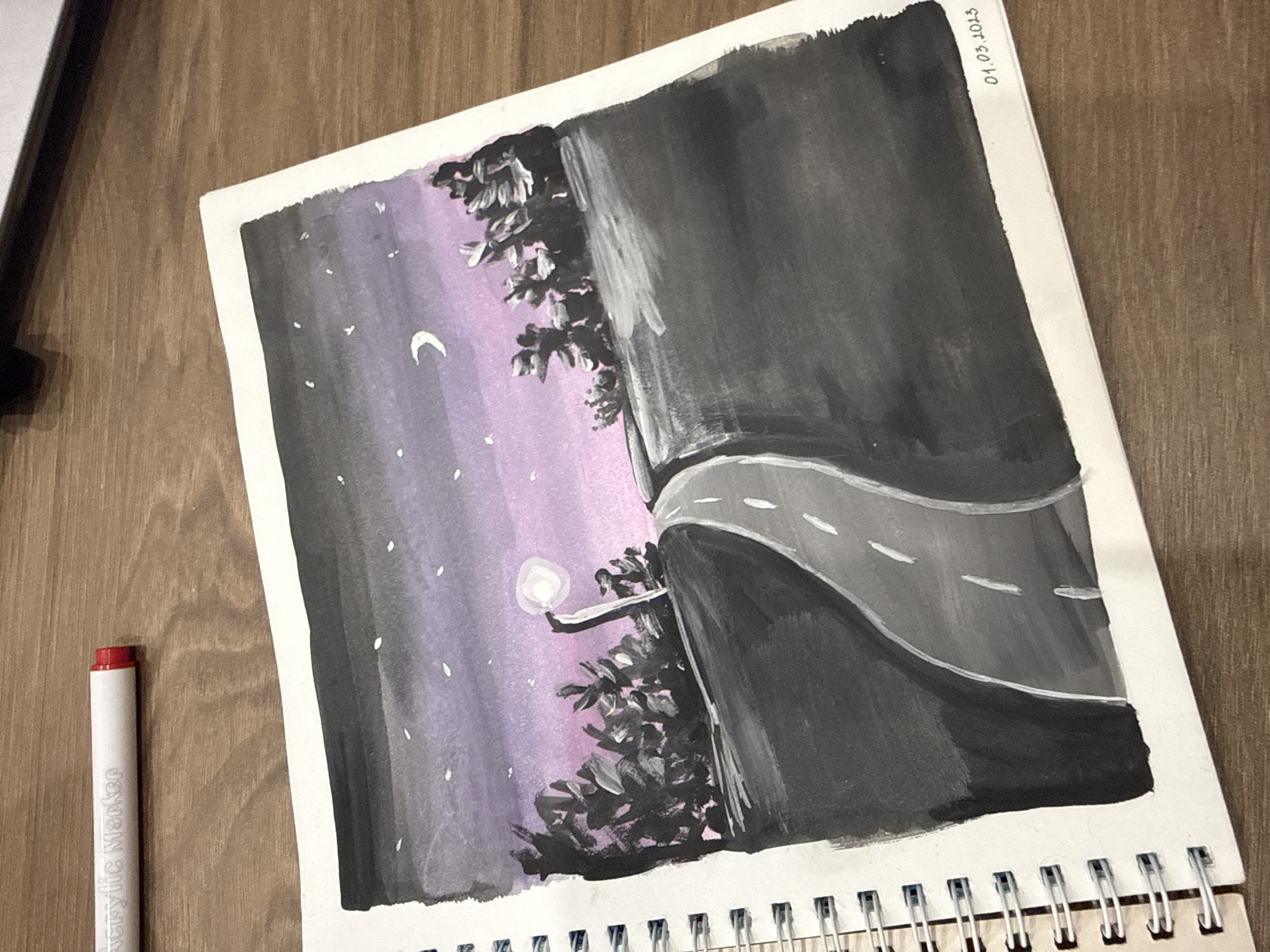 The image shows a drawing of a nighttime landscape with a road curving through dark trees under a starry, purple sky with a crescent moon. A single lamppost illuminates the foreground. The drawing is on a spiral-bound sketchpad, with the date "01.03.2023" written in the bottom right corner. A white acrylic marker with a red cap is visible in the upper left corner of the image, partially obscuring the text "Acrylic Marker" on its side. There are no people or animals depicted in the drawing. The location in the city of Murom, Russia, is not specified by any visual cues in the image.