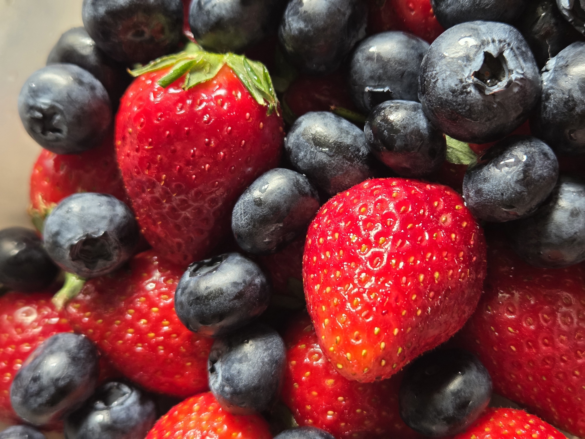 A close-up shot shows a pile of strawberries and blueberries. The strawberries are bright red with tiny yellow seeds, and some have green stems. The blueberries are a deep blue-purple color and have a smooth, waxy appearance. The berries are clustered together, with some overlapping others. The scene is brightly lit, emphasizing the textures and colors of the fruit. No location or city is discernible from the image. No people, actions, or interactions are present.