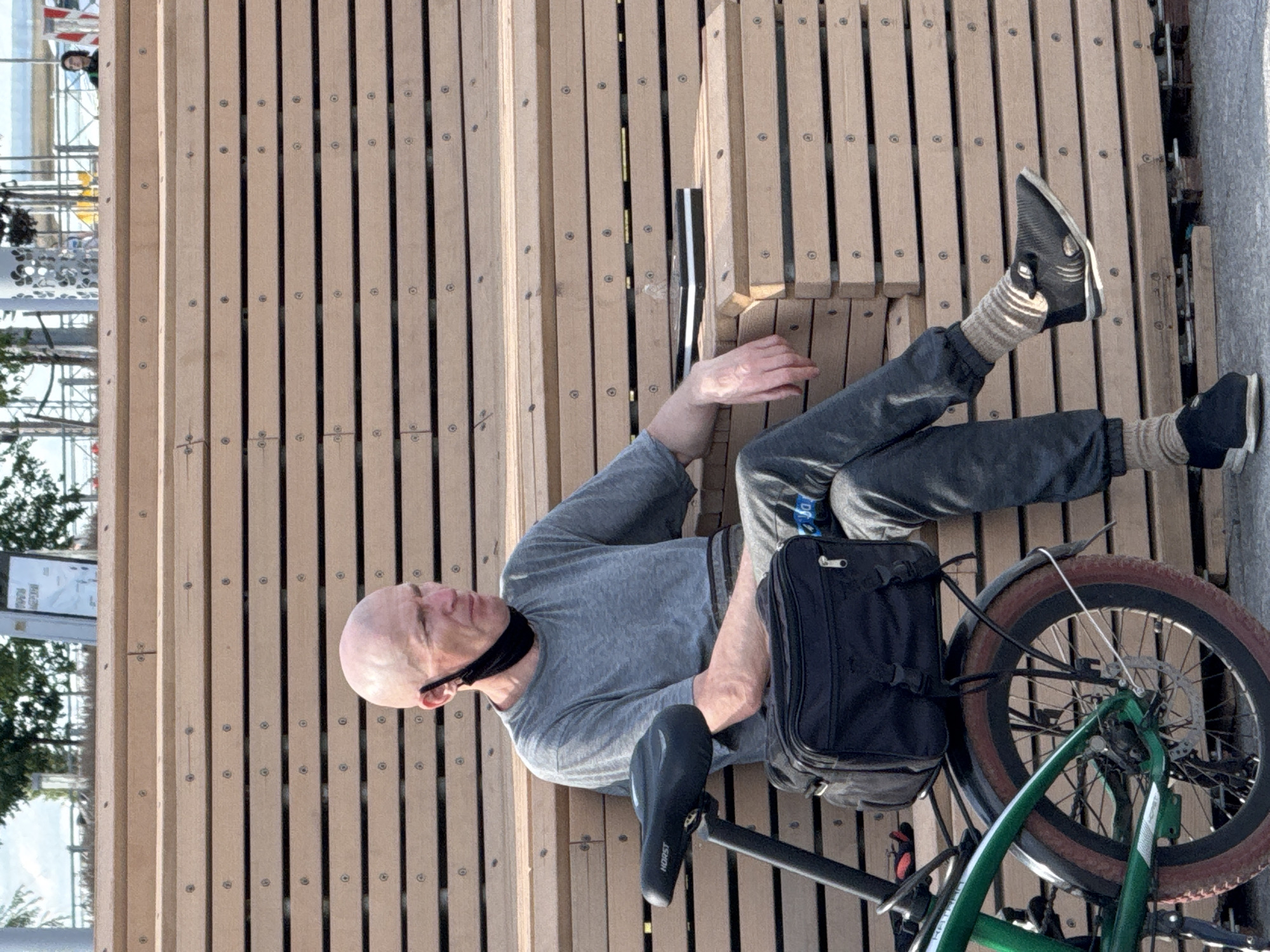 A bald Caucasian man wearing a grey t-shirt, grey sweatpants, and striped socks is sitting on a wooden bench in Saint Petersburg, Russia. He has a black mask hanging from his right ear. A black bag is attached to the back of a green bicycle next to him. The man is looking to his right. A laptop is resting on the bench to his right. The wooden bench is made of horizontal slats. The background shows trees and buildings.