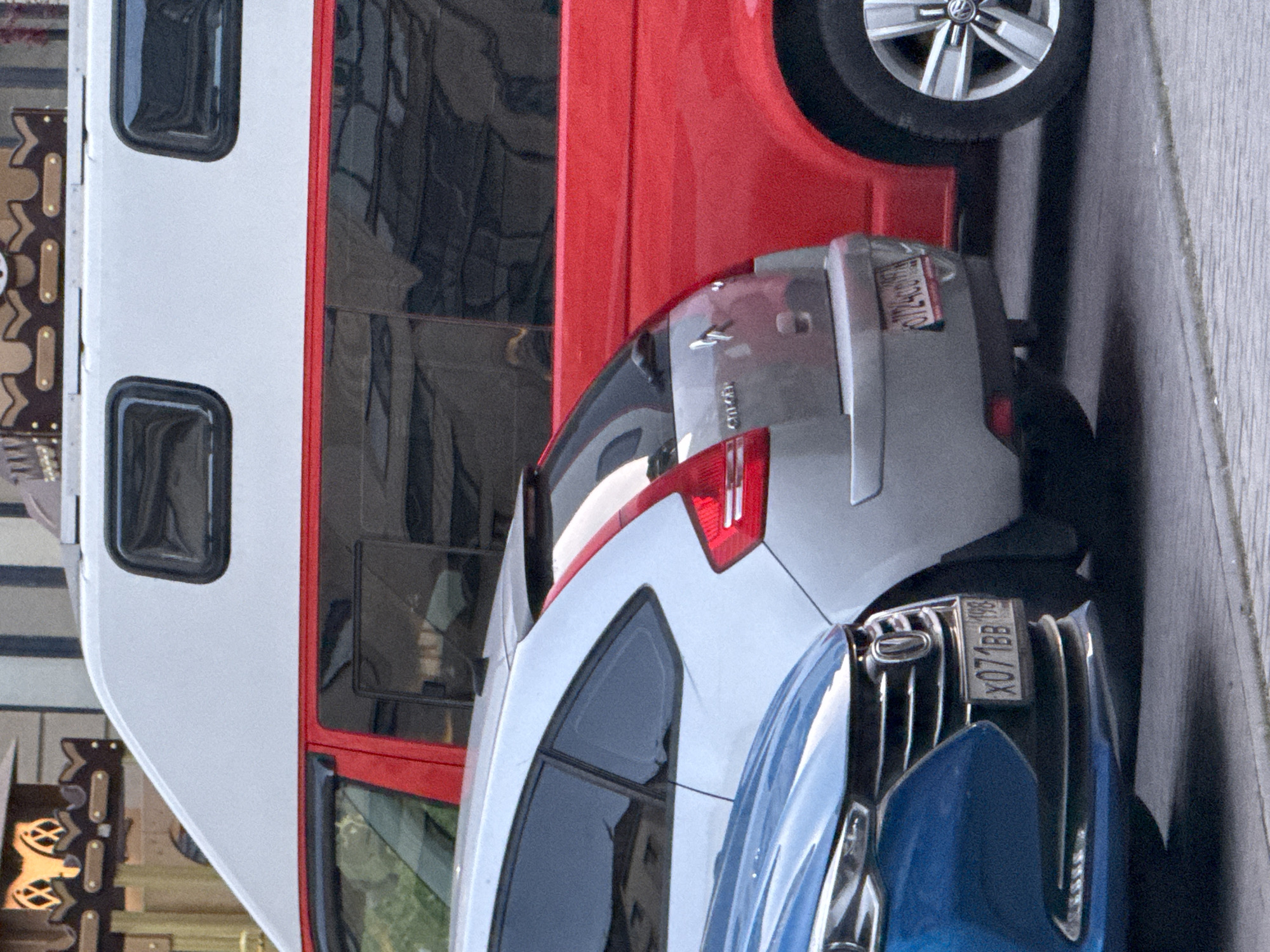 The image shows a close-up of several parked vehicles in what appears to be a street setting. The license plates indicate the location is likely Zelenogradsk, Russia. From left to right, there's a white van with a red stripe, part of a red vehicle above a grey car and a blue car, partially visible and closest to the viewer. The vehicles are tightly packed. The focus is on the rear portions of the cars, including taillights and license plates. The environment suggests a city street with pedestrian walkways.