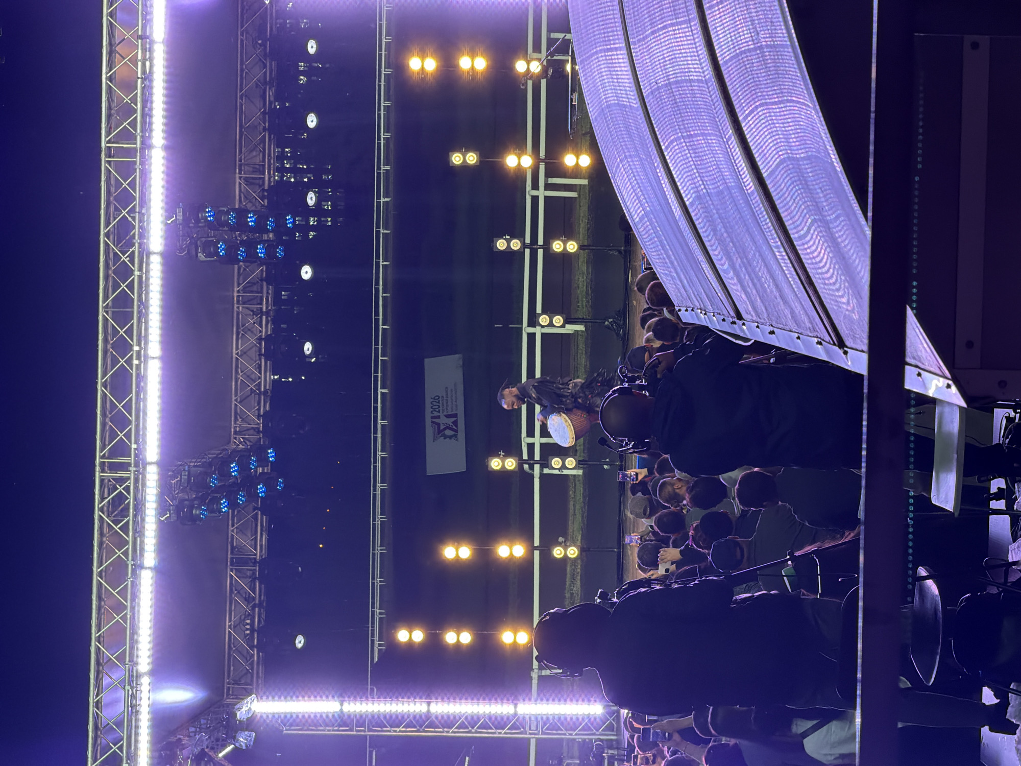 The image depicts a nighttime outdoor event with a stage and a crowd. Several people are visible in the foreground, some facing away from the camera towards the stage, and others facing towards the stage, some holding up phones. A performer is on stage, playing a large drum. The stage is lit with bright purple and yellow lights, and there are large video screens or panels in the foreground, illuminated by purple lights. The background appears to be a dark, open area, possibly a field or track, with a white fence. The event seems to be well-attended, with a significant number of people gathered to watch the performance.