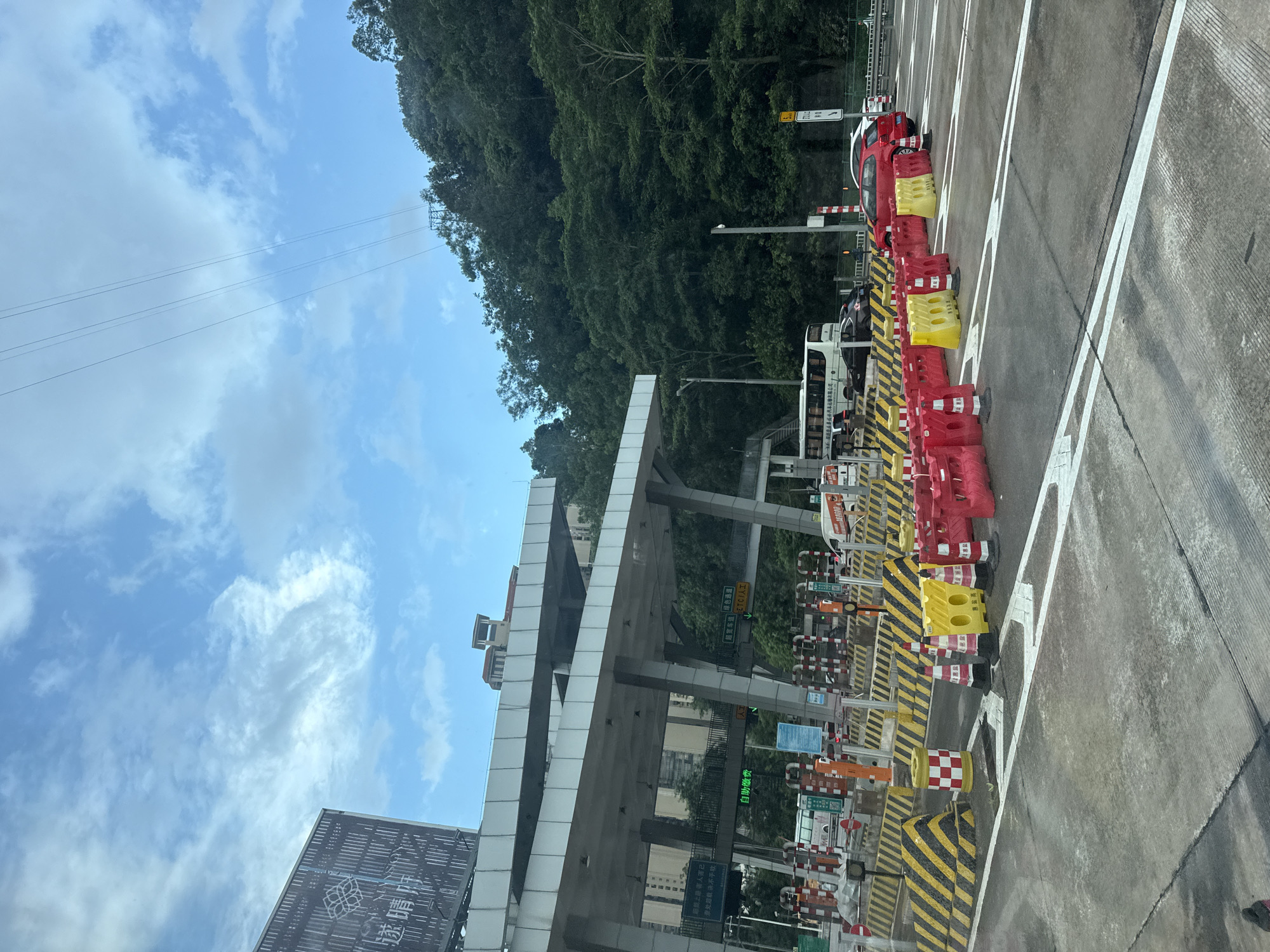 The image shows a toll station on a highway in Huizhou, China, under a partly cloudy sky. The toll station has multiple lanes, with some open and others closed off by red and yellow barriers and traffic cones. Cars and a bus are visible in the lanes. The toll station structure is large and modern, with white canopies supported by pillars. In the background, there are buildings and a densely forested hillside. Power lines are strung across the sky. The road surface is asphalt, with white lane markings. The overall scene depicts a busy transportation hub in a natural setting.