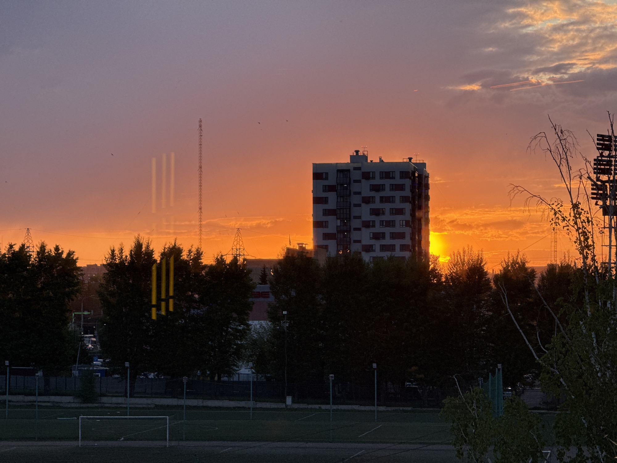 A photograph depicts a sunset over a cityscape in Krasnoyarsk, Russia. The sky is filled with hues of orange and purple, with clouds scattered throughout. Silhouetted in the foreground are trees, a soccer field with a goalpost, and a tall building. A transmission tower is visible to the left of the building. The sun is setting behind the building and trees. In the right corner, a tree with sparse branches is visible.