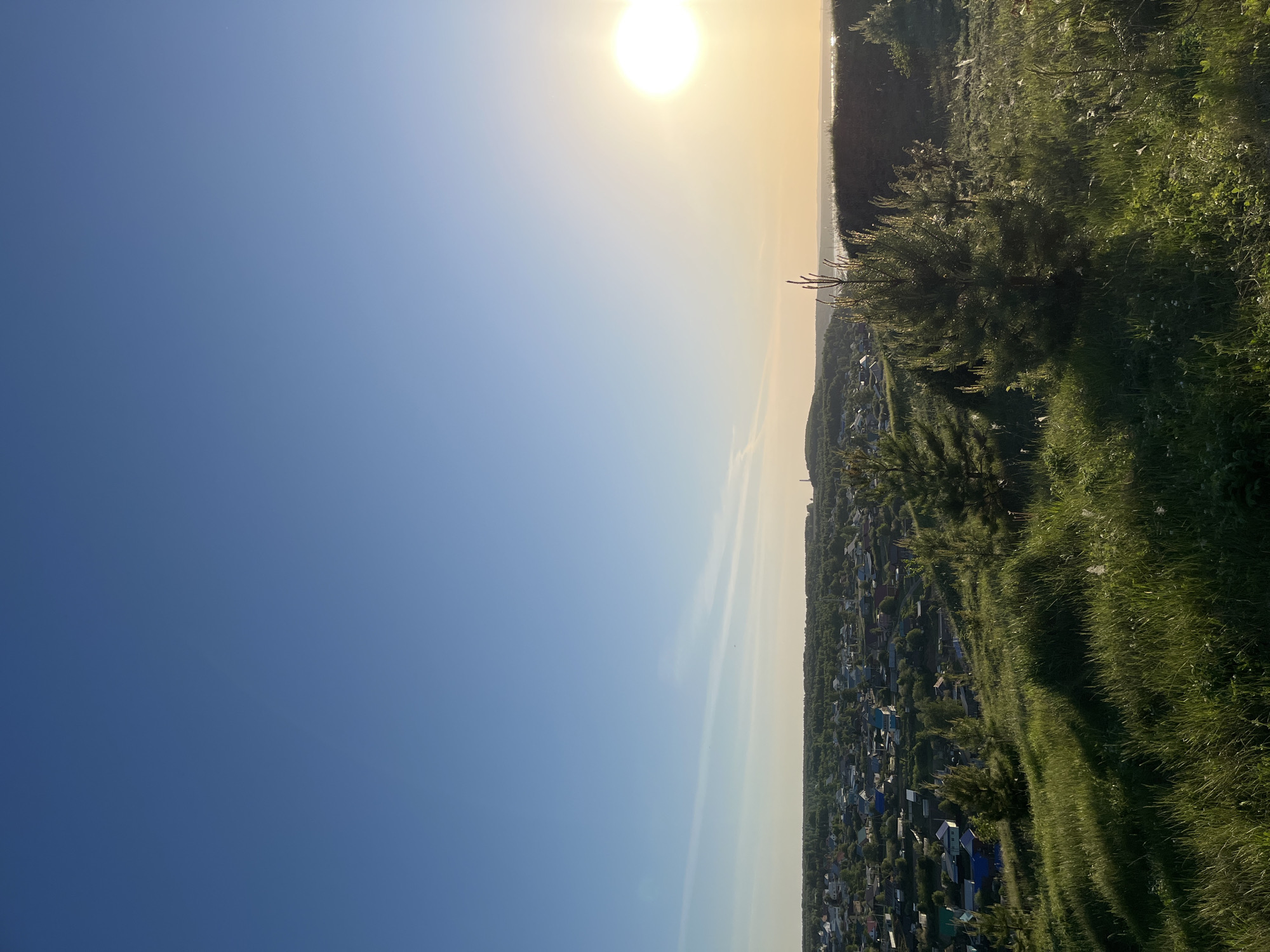 The image depicts an elevated view of Ольховка, Russia, at sunset. The sky is a gradient of blue to yellow near the sun. Below, a dense residential area stretches towards the horizon, interspersed with trees. In the foreground, there is a grassy hillside.