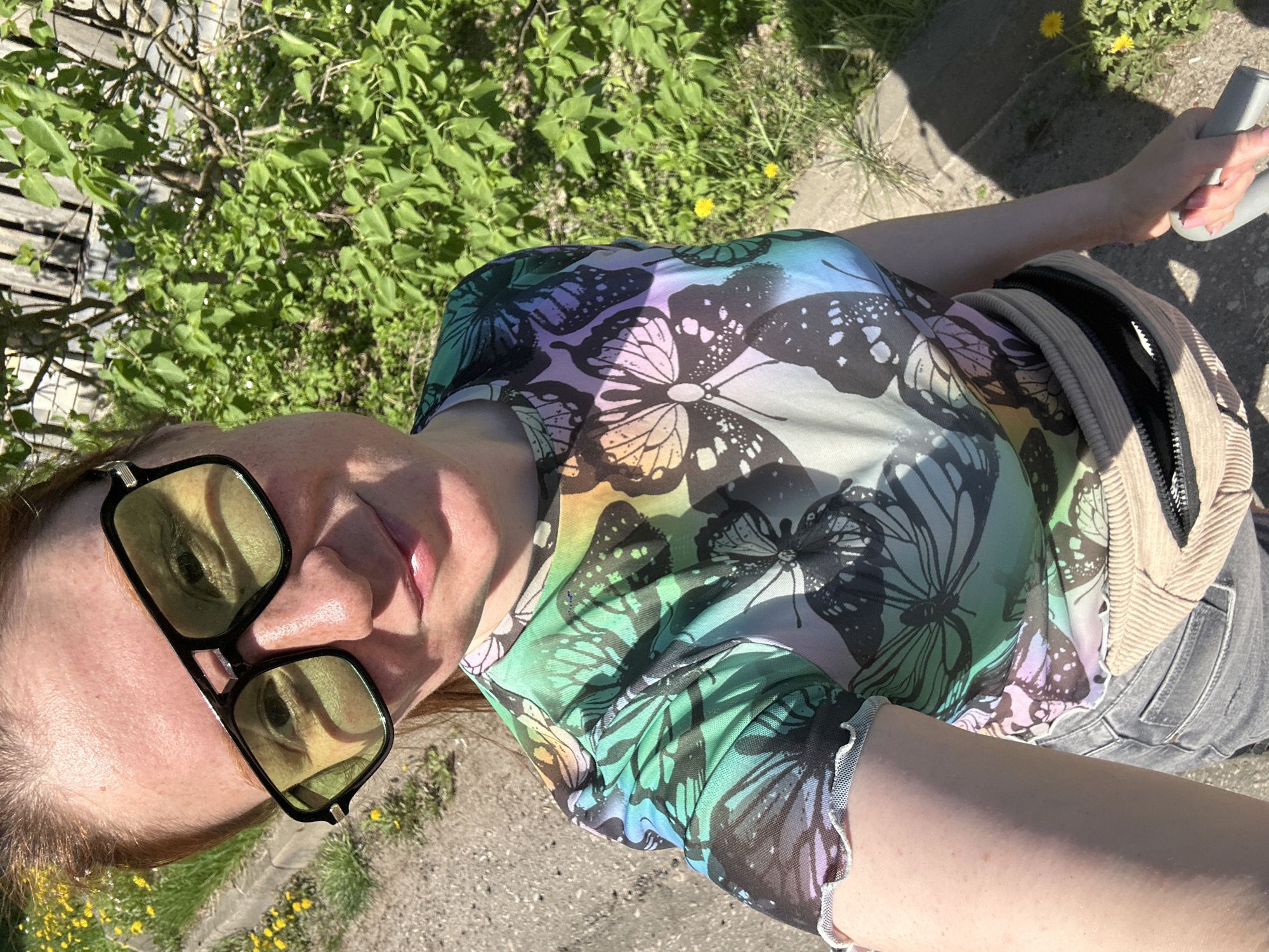 A woman with auburn hair and wearing large, square yellow sunglasses is taking a selfie outdoors. She is wearing a colorful top printed with butterfly designs. A fanny pack or small bag is slung around her waist, and she appears to be wearing gray jeans. In the background, there is greenery, including a wooden fence and dandelions on the ground. The location appears to be in a park or green space, possibly in the city of Nizhny Novgorod, Russia. She is holding onto a light gray handle, which may be part of outdoor exercise equipment.