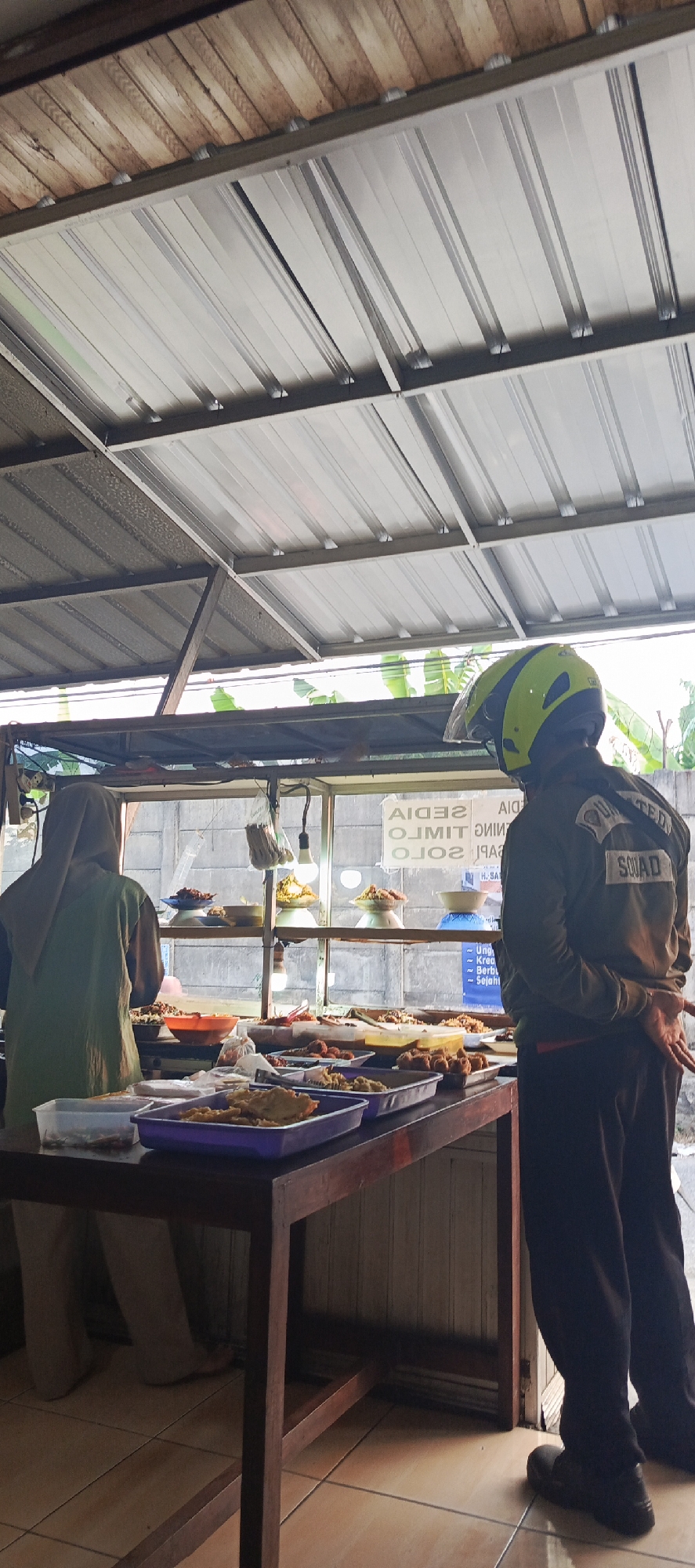 The image shows a food stall in Sukaresmi, Indonesia. A person wearing a hijab and an olive-green tunic is standing behind a wooden counter, facing away from the camera and towards a glass display case filled with various food items. Another person, wearing a dark jacket with "SQUAD" written on the back in white lettering and a neon yellow helmet, is standing to the right, also facing the food display. The food stall has a corrugated metal roof supported by metal beams. The background shows a textured wall and some foliage. The lighting appears to be natural, possibly from the overcast sky. The overall scene depicts a typical street food vendor setup.