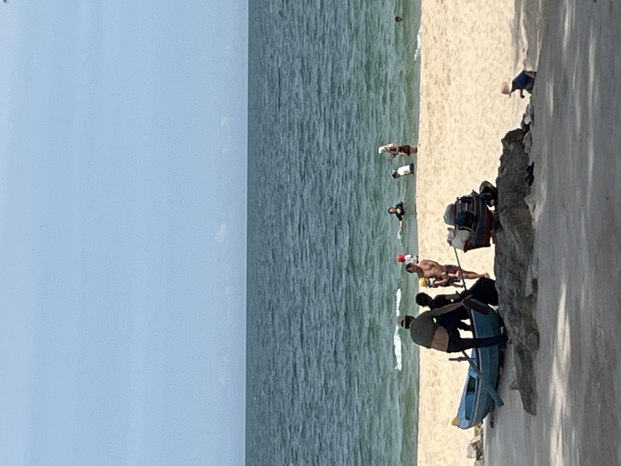 In Mũi Né, Vietnam, on a sandy beach, several people are engaged in various activities by the ocean. In the foreground, a blue fishing boat is partially on the sand. Three individuals, appearing to be adults, are gathered around the boat; one wearing a green shirt and cap, another in a dark shirt, and a third partially obscured. Nearby, a small child wearing a blue shirt and a straw hat is sitting on the sand. Further back, several people are in the shallow water: one person in a black swimsuit, another in a white t-shirt and dark shorts, and a woman in a light-colored headscarf and dark clothing. A man and a child in red swimwear are also visible. The ocean stretches out to the horizon under a clear, pale blue sky.