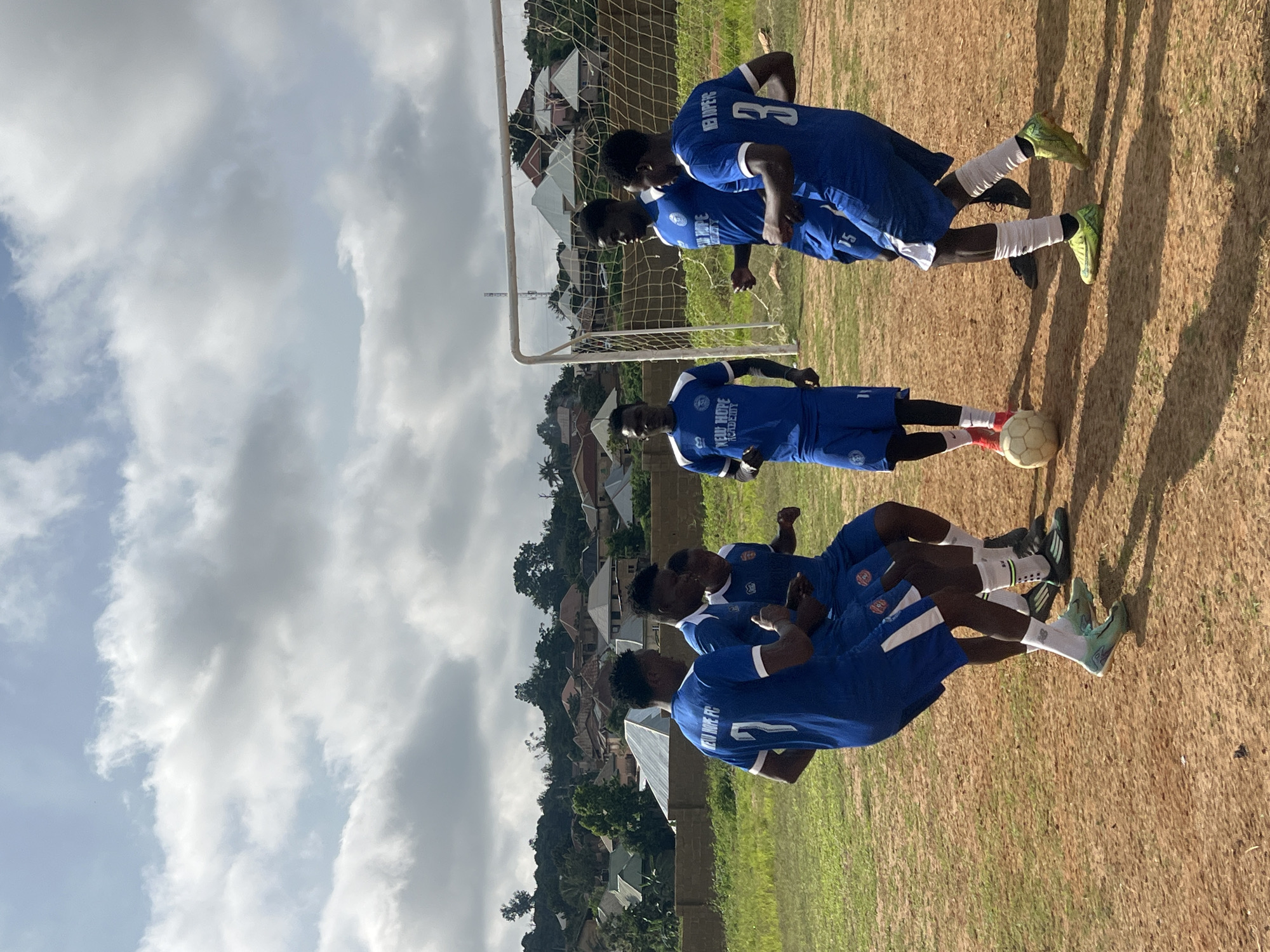 A group of five young men, all wearing blue soccer uniforms with white trim and the inscription "NEW HOPE FC ACADEMY", are gathered on a dirt soccer field in Jos, Nigeria. They are in various stages of activity around a white soccer ball. The men are of African descent, with dark skin and hair. Their uniforms consist of short-sleeved jerseys and shorts, with white socks and soccer cleats. One man in the foreground, wearing jersey number 7, is facing away from the camera. Another man, wearing jersey number 15, is in the center, looking down at the ball. Two other men are positioned on either side of him, with one wearing jersey number 3 on the right. A fifth man, wearing jersey number 11, is visible on the far left. Behind the soccer field, a chain-link soccer goal with a white net is set up. Further in the background, a densely populated residential area with various houses and buildings is visible, situated on a hillside. The sky above is partly cloudy with patches of blue sky visible. The overall impression is a casual soccer practice or gathering.