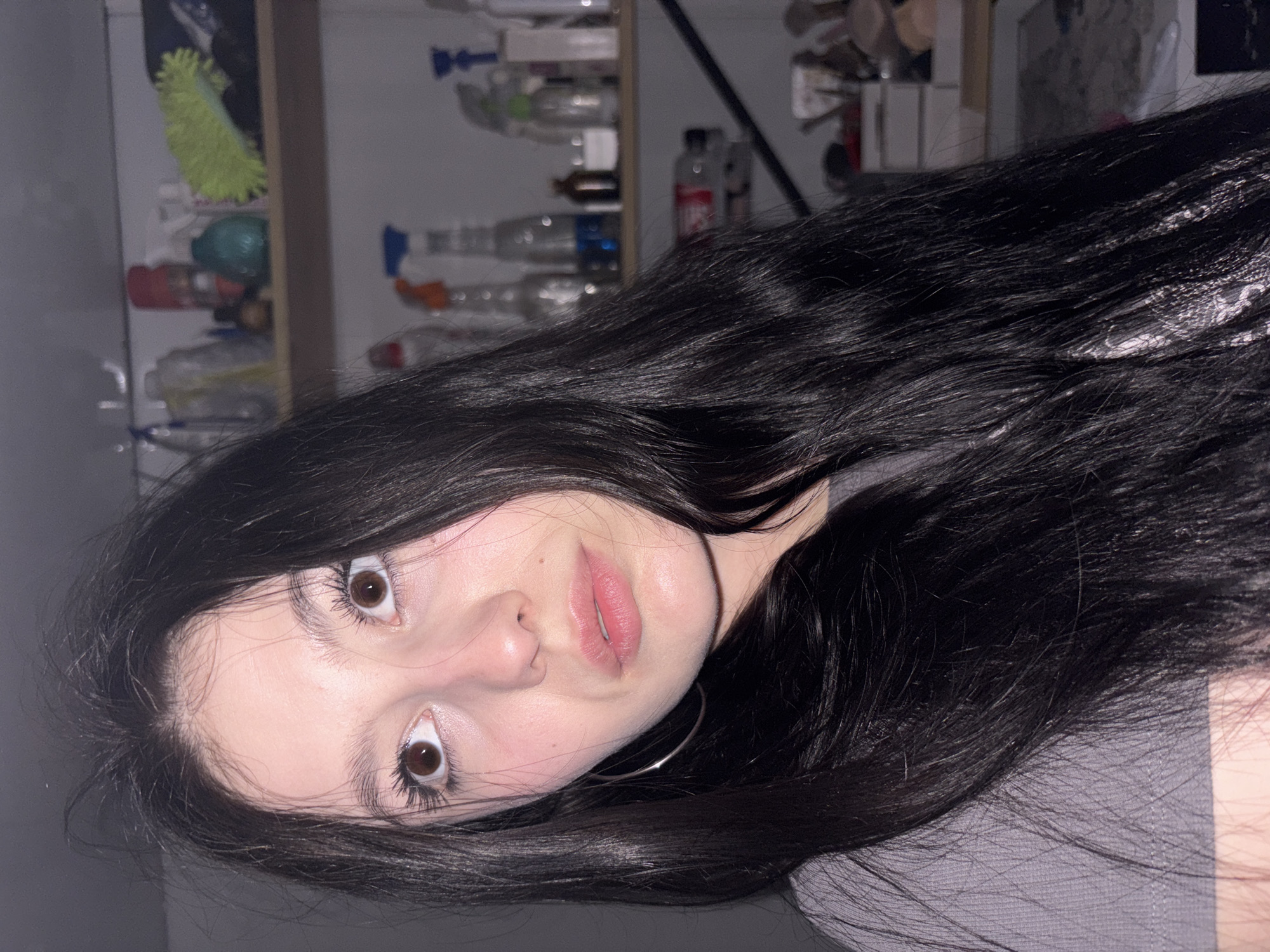 A close-up, rotated image depicts a young woman with long, dark hair cascading around her face. Her skin tone is pale, and her eyes are brown. She has full lips with a pink tint. A silver hoop earring is visible on her left side. The background shows a cluttered shelf containing various items, including cleaning supplies and toiletries. The location is likely indoors, but the image doesn't provide specific details indicating it's in Фоки, Russia.