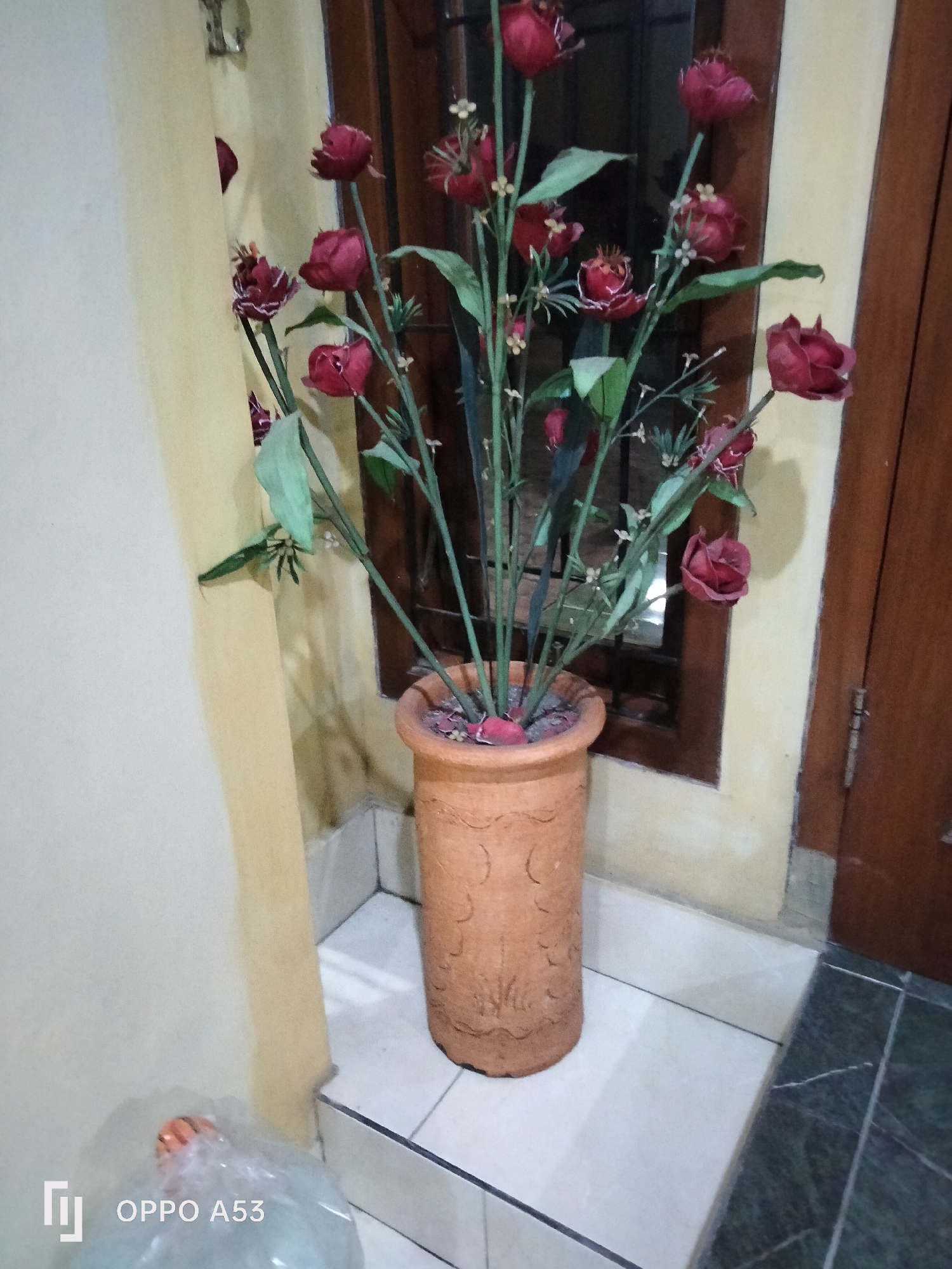 A tall, cylindrical, terracotta pot containing artificial red roses stands on a tiled floor next to a cream-colored wall and a wooden door. The pot is decorated with embossed patterns. The artificial roses have dark red petals and green stems. Behind the pot, a window with dark brown trim is visible. To the left of the pot, a plastic bag containing an orange object is on the floor. The scene appears to be indoors, possibly in a residential setting in Rejowinangun, Indonesia.