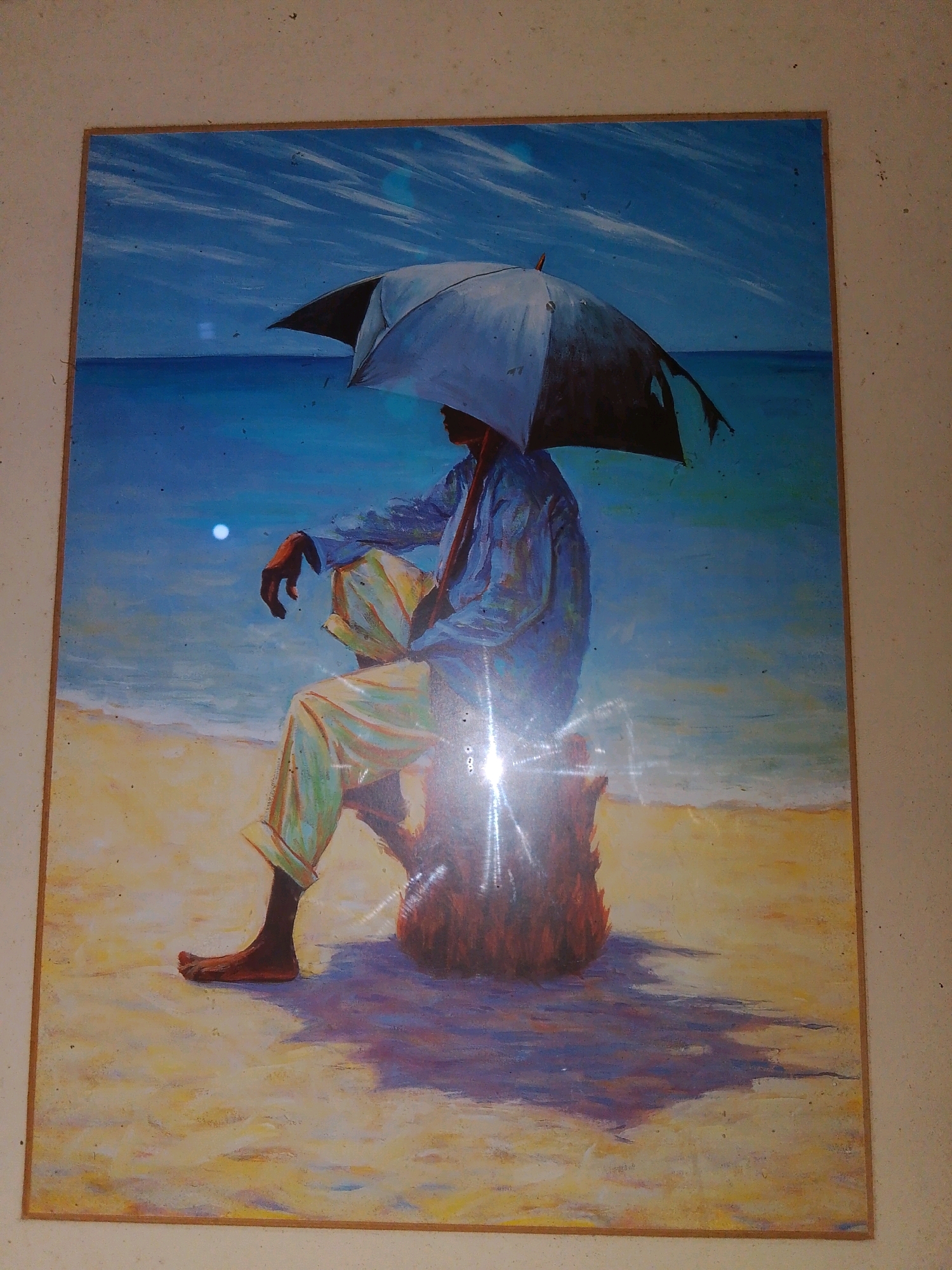 This is a painting of a person sitting on a beach under an umbrella. The person is wearing a blue shirt and light-colored pants. They are sitting on a reddish-brown object, possibly a log or rock, with their legs crossed. The background is a blue sky with white clouds and a calm blue ocean. The foreground is a sandy beach with shadows cast by the person and the umbrella. There is a bright reflection on the surface of the painting. The painting is framed with a thin brown border. The image shows no indication of the city of Junda ward, Kenya.