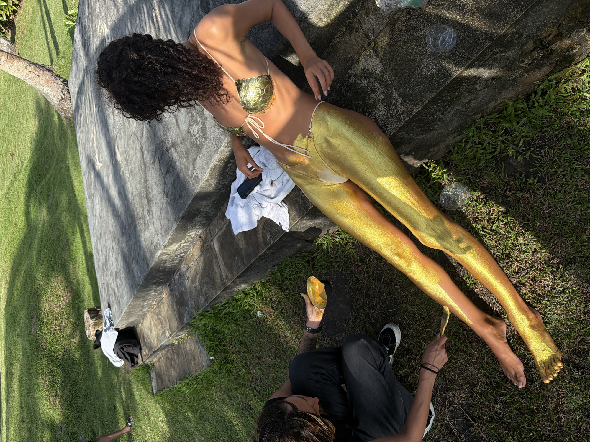 A woman with curly hair lies on a concrete structure in Kerambitan, Indonesia. She wears a gold bikini top and gold paint covers the majority of her body. Another person sits on the grass, applying gold paint to her legs with a brush. A white towel and a black object rest nearby. Clear plastic spheres are scattered on the grass near the concrete structure. The surrounding environment consists of a grassy lawn and a tree trunk is visible in the background.