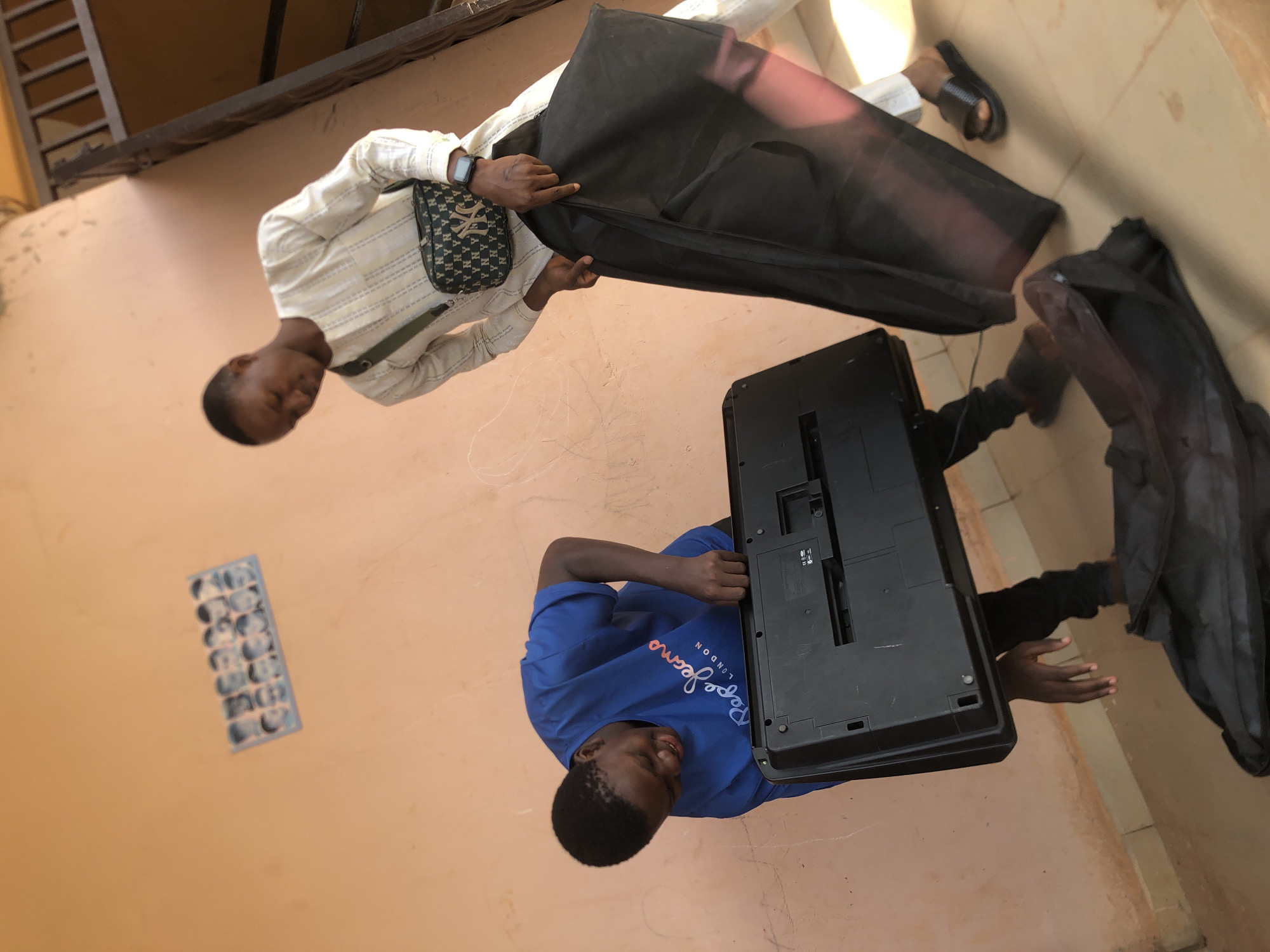 The image shows two young men in Nkwelle Ezunaka, Nigeria. One man, wearing a striped long-sleeved shirt and a patterned cross-body bag, is holding open a black case. The other man, wearing a blue t-shirt, is holding a black electronic keyboard. They are outdoors on a light orange-colored tiled surface next to a wall of the same color. A window with a grid of small panes is visible on the wall.