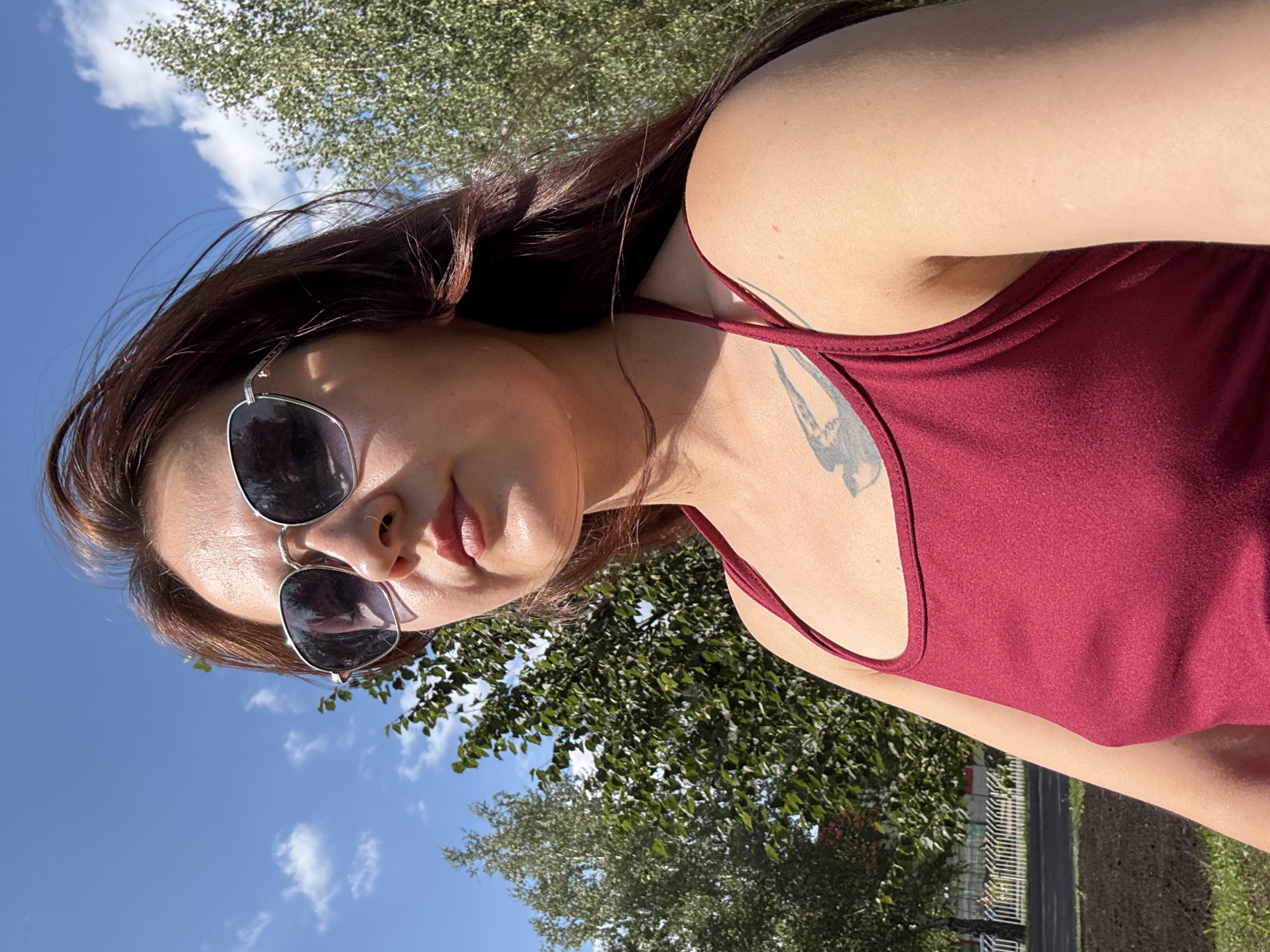 A woman wearing sunglasses and a red tank top is outdoors on a sunny day. She has brown hair and a tattoo on her chest. She is looking up and to the left. In the background, there are trees and a blue sky with clouds. The location is specified as Naberezhnye Chelny, Russia.