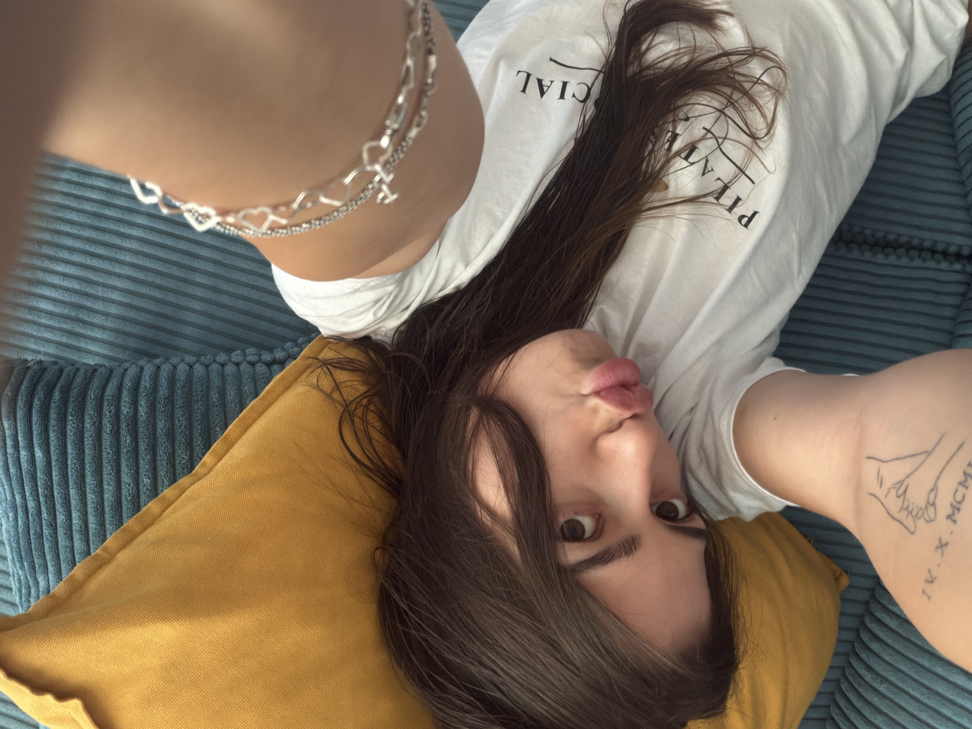 In this overhead shot, a young woman lies on a teal corduroy couch with a mustard yellow pillow supporting her head. She is wearing a white t-shirt and a silver bracelet with heart charms adorns her left wrist. Her dark brown hair is spread across her face and the pillow. The woman is looking at the camera with wide eyes and her lips are pursed. A tattoo is visible on her right arm, featuring two hands clasped together and Roman numerals. The lighting is soft and natural, suggesting an indoor setting.