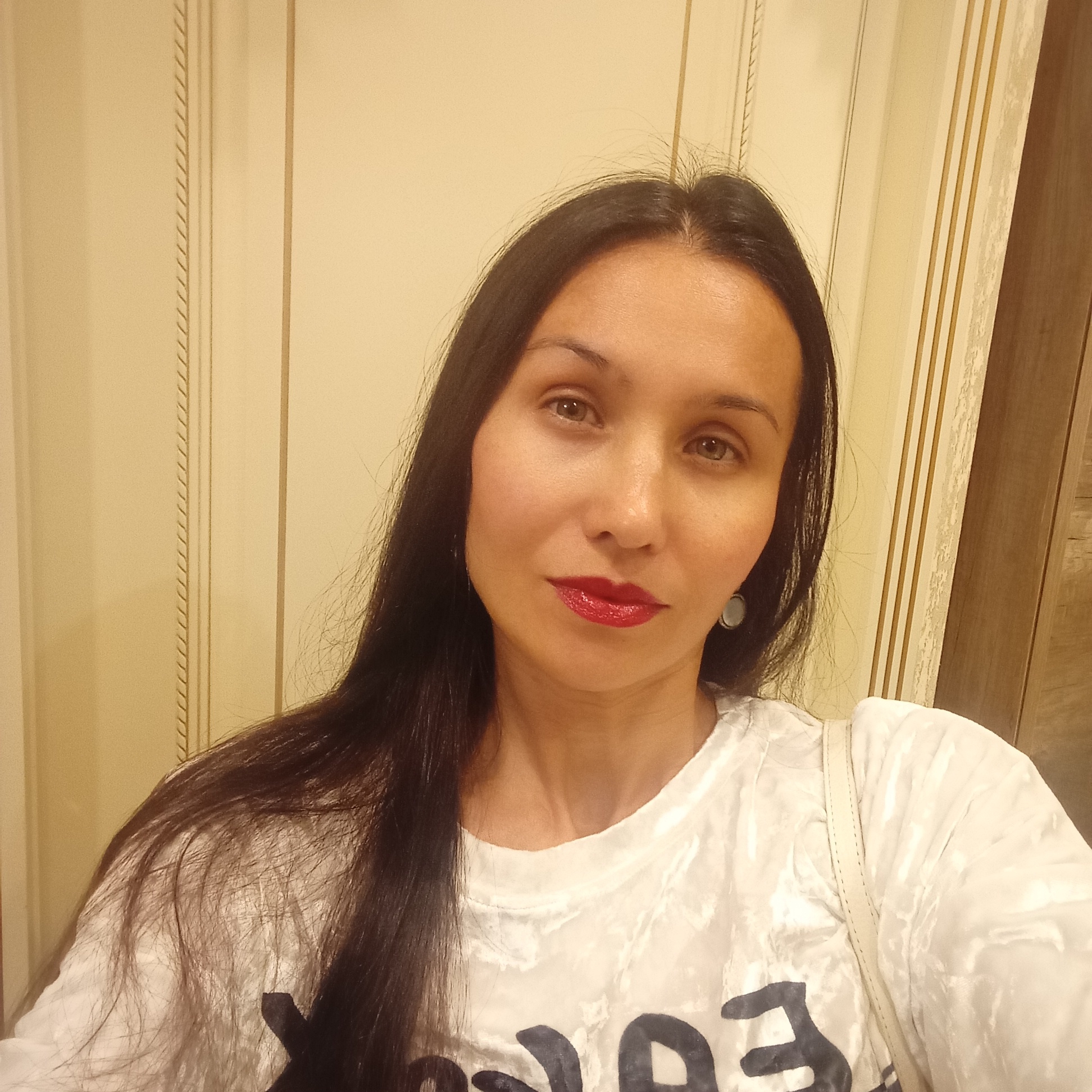 A woman with long, dark hair and fair skin takes a selfie. She is wearing a white t-shirt and red lipstick. Her eyes are light brown and she has a faint smile. The background appears to be a wall with vertical paneling and decorative trim, suggesting an indoor setting. The lighting is warm and appears to be artificial. There is no visible indication of the city of Sochi, Russia.