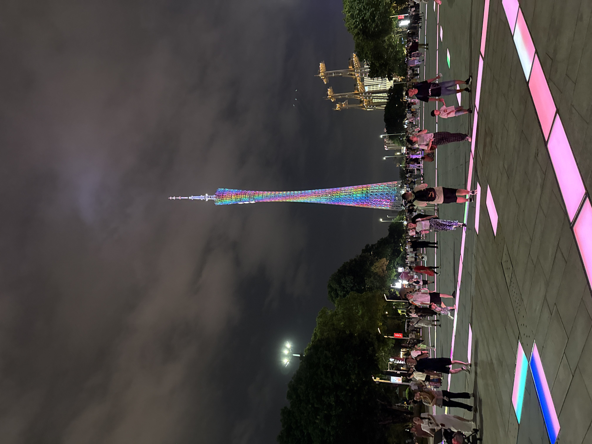 The image depicts a nighttime scene in Tianhe District, China, featuring the Canton Tower prominently in the background. The tower is illuminated with a vibrant rainbow of colors, creating a striking visual against the dark, cloudy sky. In the foreground, a group of people are gathered on a paved plaza, which is illuminated by colorful, embedded lights. The people are casually dressed and appear to be enjoying the evening. Some are walking, while others are standing and observing the tower or interacting with each other. The presence of streetlights and the illuminated buildings in the distance suggest an urban environment. The overall atmosphere seems relaxed and festive, with the colorful lights contributing to a celebratory mood. The time of day is clearly night, and the cloudy sky adds a dramatic effect to the scene.