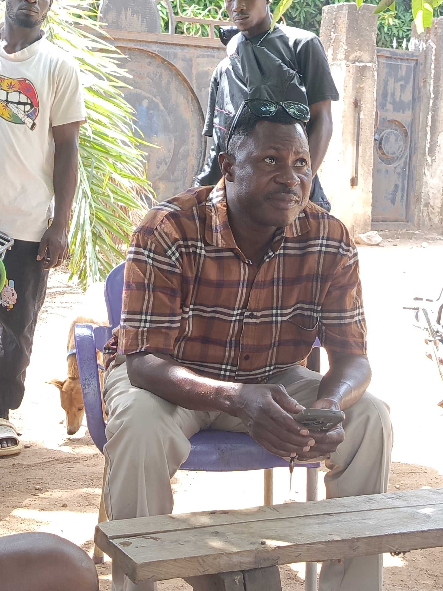 The image was taken outdoors in Akwanga, Nigeria. A man is seated on a purple plastic chair, holding a mobile phone. He is wearing a brown plaid shirt and khaki pants. Sunglasses are resting on his head. In the background, there are three people standing. A dog is under the chair. In front of the seated man, there is a weathered wooden table. A closed gate and other outdoor structures are visible.