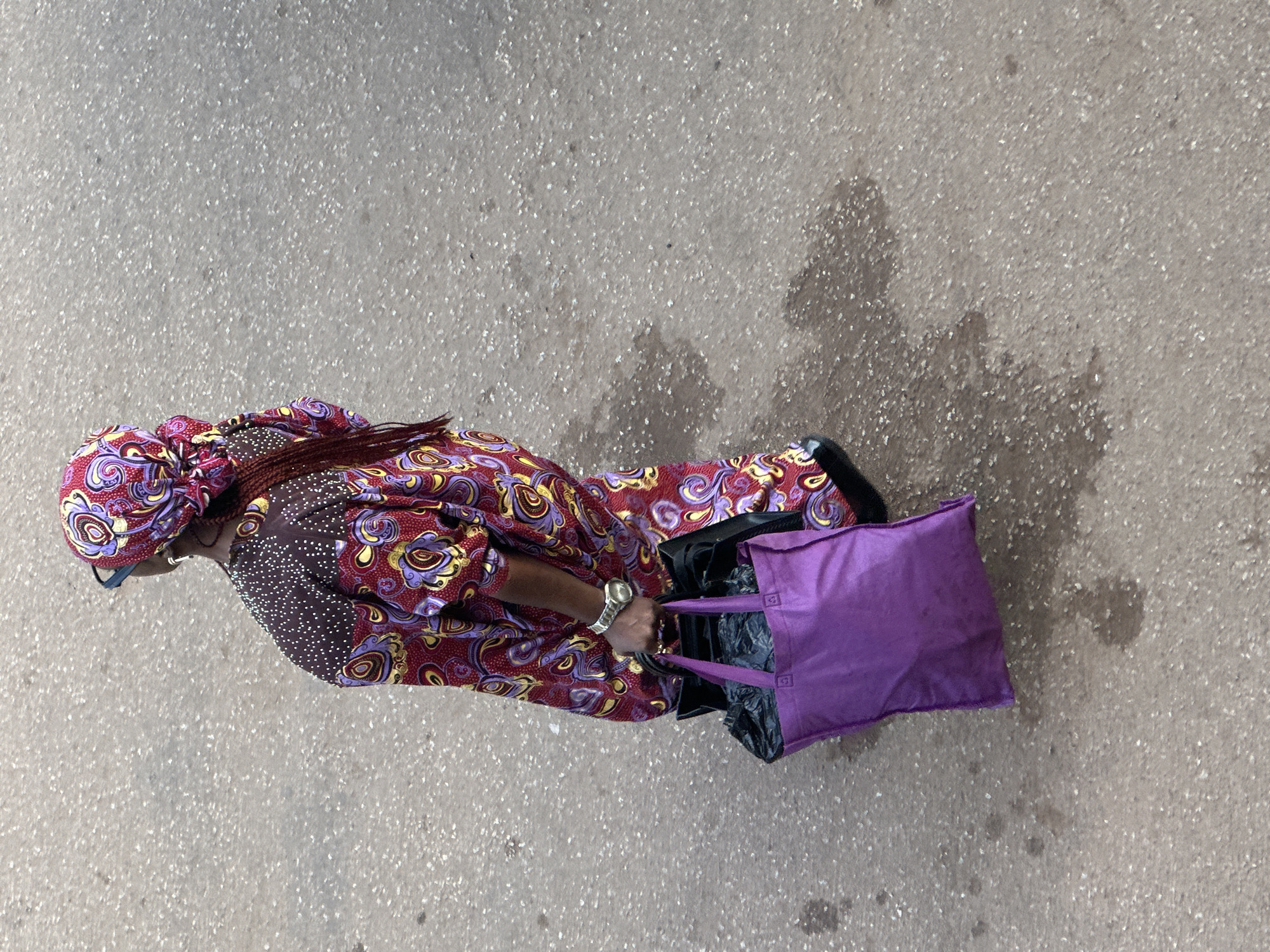 An overhead, high-angle shot shows a person walking on a paved surface in Bukuru, Nigeria. The person is wearing a red and purple patterned garment with gold accents on the shoulders and a matching headscarf. They are also wearing blue-tinted glasses and a silver watch on their left wrist. The person is carrying two bags in their left hand: a black bag partially obscured by a bright purple tote bag. The ground is a speckled, grey asphalt surface with some damp patches. The scene is brightly lit, suggesting daylight.