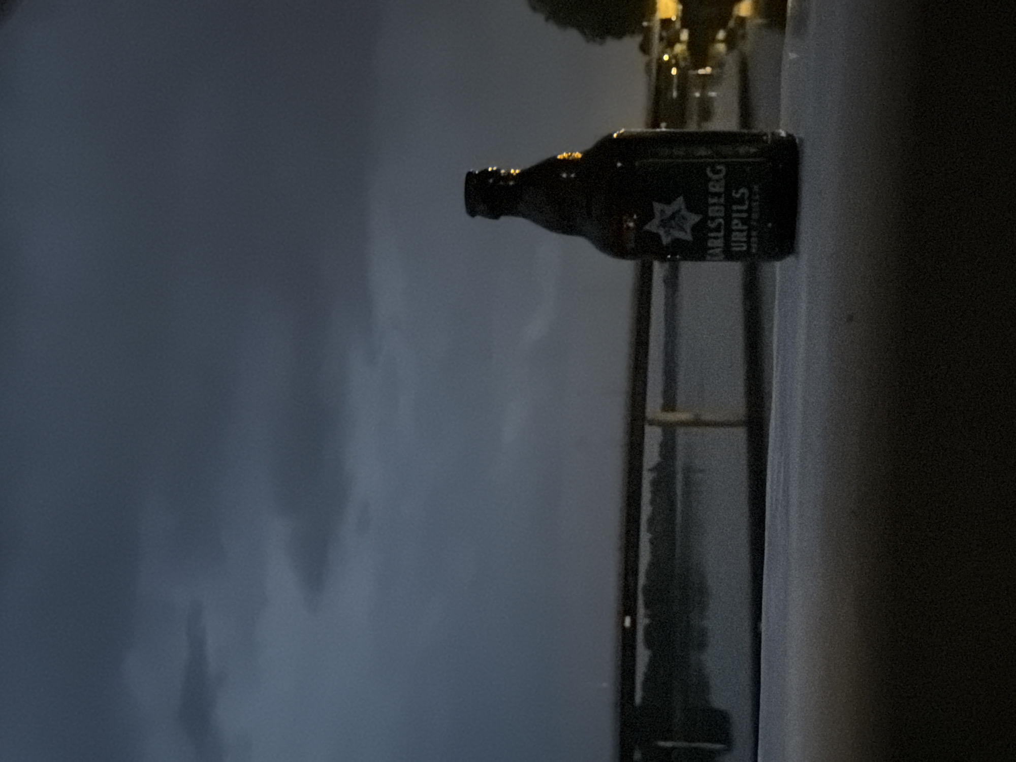 In Charnay-lès-Mâcon, France, at twilight, a brown glass bottle of Carlsberg Urpils beer stands on a dark, textured surface, likely a table or railing, in the foreground. The bottle is partially illuminated, with a label displaying the Carlsberg logo and text. Behind the bottle, a body of water, possibly a river, reflects the dim sky. A dark, horizontal structure, resembling a bridge or pier, spans across the water in the mid-ground. The background features a silhouette of trees and distant lights from buildings on the far bank. The sky above is overcast with dark, heavy clouds, suggesting impending rain or a storm. The overall atmosphere is moody and somber, with low light conditions and a muted color palette.