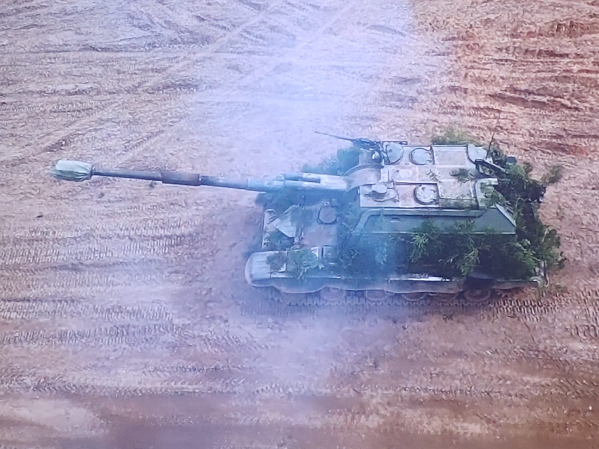 An aerial, slightly hazy, medium shot captures a camouflaged military tank in a dirt field. The tank is positioned diagonally across the frame, facing left. Its hull is a muted green and tan, partially obscured by foliage, giving it a camouflaged appearance. The tank's turret is visible, with what appears to be a machine gun mounted on top. A long barrel extends forward from the turret, ending in a covered muzzle. The ground beneath the tank is a dusty, textured surface with visible tire tracks or tread marks. The surrounding environment is a flat, open area with a similar dirt texture. The image does not contain any information to suggest a specific location in Molenbeek-Saint-Jean - Sint-Jans-Molenbeek, Belgium, nor does it depict any people or specific actions.