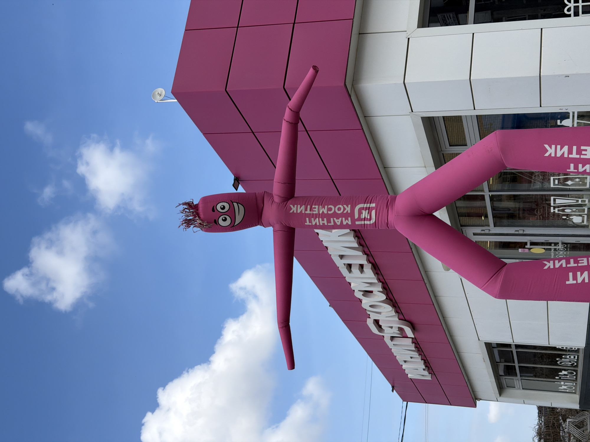 A tall, pink inflatable air dancer, branded with the white text "МАГНИТ КОСМЕТИК" (Magnit Kosmetik) and the Magnit logo, stands in front of a commercial building. The inflatable figure features a cartoonish face with prominent eyes and an open mouth, with spiky extensions on its head, and its arms are extended horizontally. The building, identified as a Magnit Kosmetik store in Krasnodar, Russia, is clad in a combination of pink and white panels. A large, three-dimensional white sign displaying "МАГНИТ КОСМЕТИК" is mounted on the upper pink section of the facade. The lower portion of the building exhibits large glass windows. A segment of the building's roof, featuring a small, white dish antenna, is visible against a clear blue sky with scattered white clouds. Overhead power lines are present on the left side of the frame. No human subjects are visible.