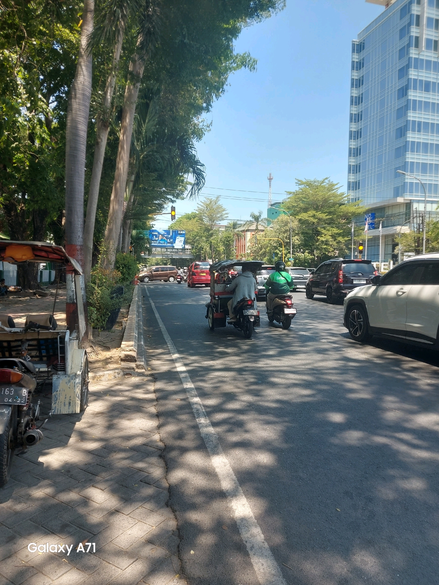 The image depicts a sunny daytime scene on a city street in Makassar, Indonesia. The foreground features a sidewalk paved with interlocking concrete blocks, with shadows from trees falling on it. A parked three-wheeled vehicle, resembling a modified motorcycle with a passenger cabin, is visible on the left. Its license plate reads "163 B 04-22".
The street is asphalt and appears to be a multi-lane road with traffic. Several vehicles are present, including a white SUV, a dark SUV, a red car, and a brown car. Two motorcycles are on the road; one is being ridden by a person wearing a green jacket with the "Grab" logo, and another is a three-wheeled motorcycle taxi with passengers. A traffic light with a yellow light illuminated is visible in the distance.
On the right side of the street, a modern, tall building with a glass facade dominates the skyline, likely a commercial or office building. Trees line the left side of the road, providing shade. A large billboard is visible in the background, featuring an advertisement with people and text. The sky is clear and blue. The overall impression is a bustling urban environment in Makassar.