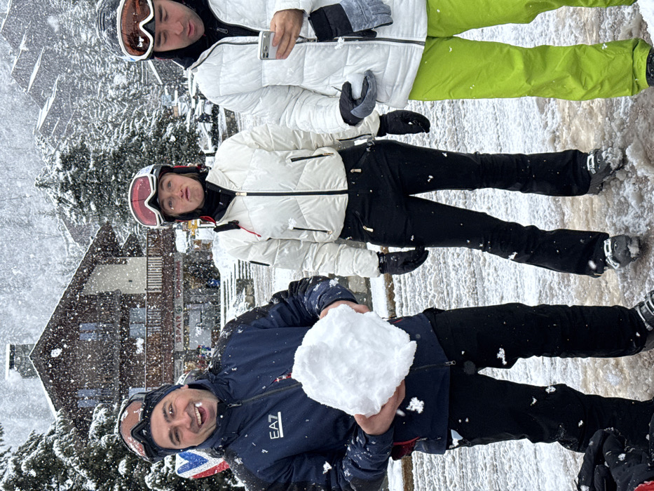 Three individuals in ski attire are depicted outdoors during active snowfall. The scene is set on a snow-covered path in a ski resort, with a chalet-style building displaying a "SPAR" grocery store sign in the background, identifying the location as Les Allues, France.
From left to right:
1. A male in a dark blue ski jacket with an "EA7" logo, dark ski pants, and a dark beanie with goggles positioned on his forehead. He is smiling broadly with an open mouth and holds a large, irregular chunk of snow with both hands.
2. A female wearing a white puffer ski jacket, black ski pants, and a white ski helmet with red goggles resting on it. Her face exhibits a puckered expression.
3. A male dressed in a white puffer ski jacket, neon green ski pants, and a black ski helmet with goggles resting on it. He holds a white smartphone in his left hand, looking down at the screen, and a small snowball in his right gloved hand.
Snowflakes are actively falling and visible across the foreground and on the subjects' clothing. The ground is covered in a mixture of packed and fresh snow. Snow-laden evergreen trees and other chalet-style buildings are present in the background.