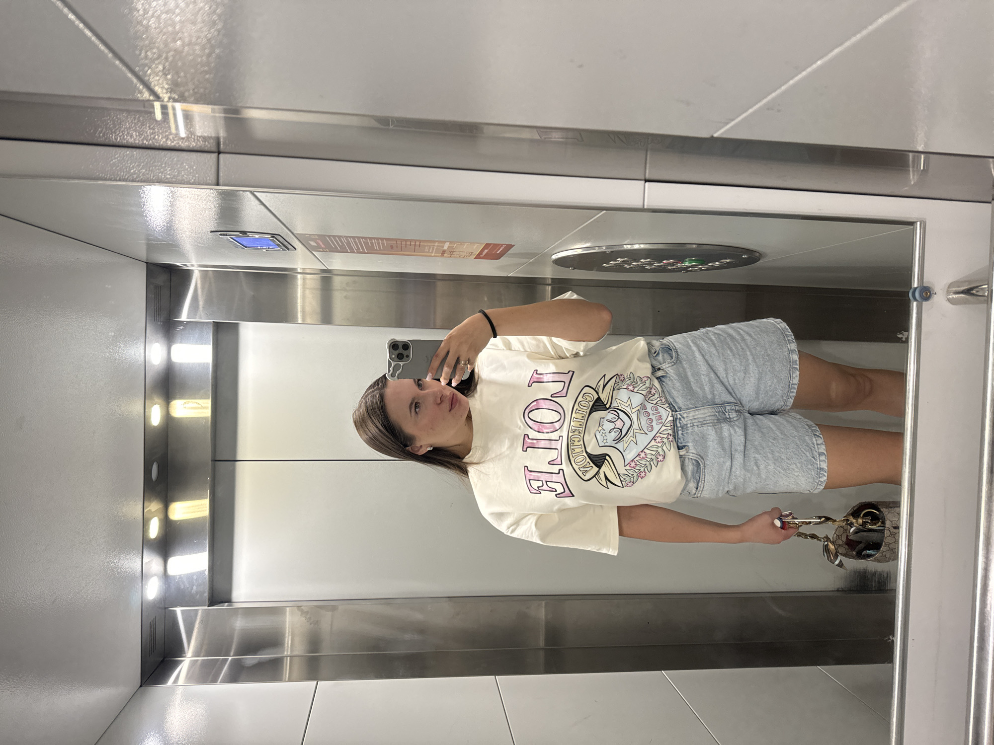 A woman is taking a selfie in an elevator. She is wearing a white t-shirt with a "LOLE" design, light blue denim shorts, and a black bracelet on her left wrist. She holds a beige and brown patterned handbag. The elevator interior is stainless steel, with recessed lighting on the left wall. The elevator is located in Akulovo, Gp Kubinka, Russia.
