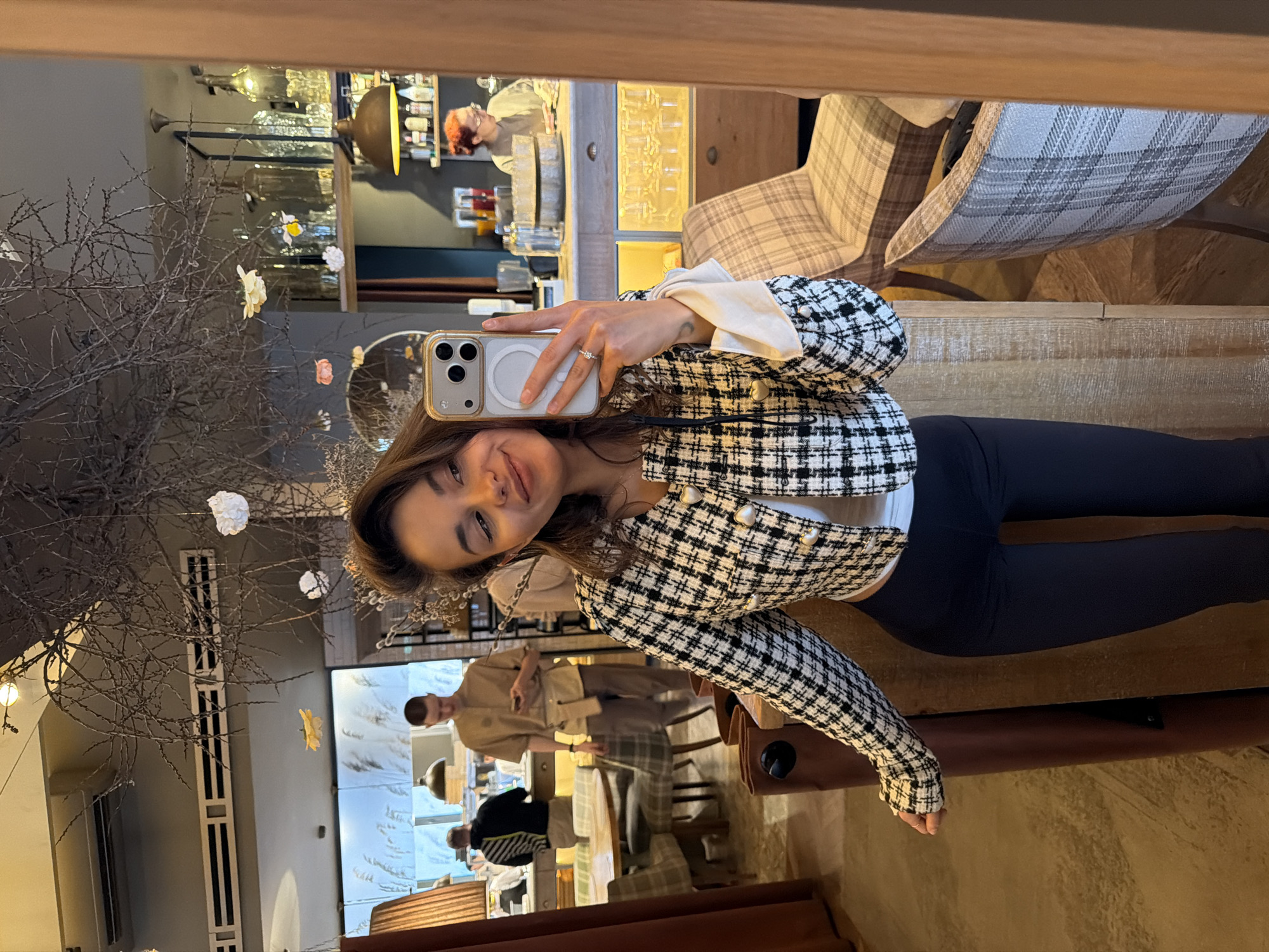 A woman is taking a selfie in a mirror. She has long brown hair and is wearing a black and white checkered jacket and black leggings. The background shows a restaurant. There are other people in the restaurant, including a bartender. The restaurant appears to be in Krasnoyarsk, Russia.