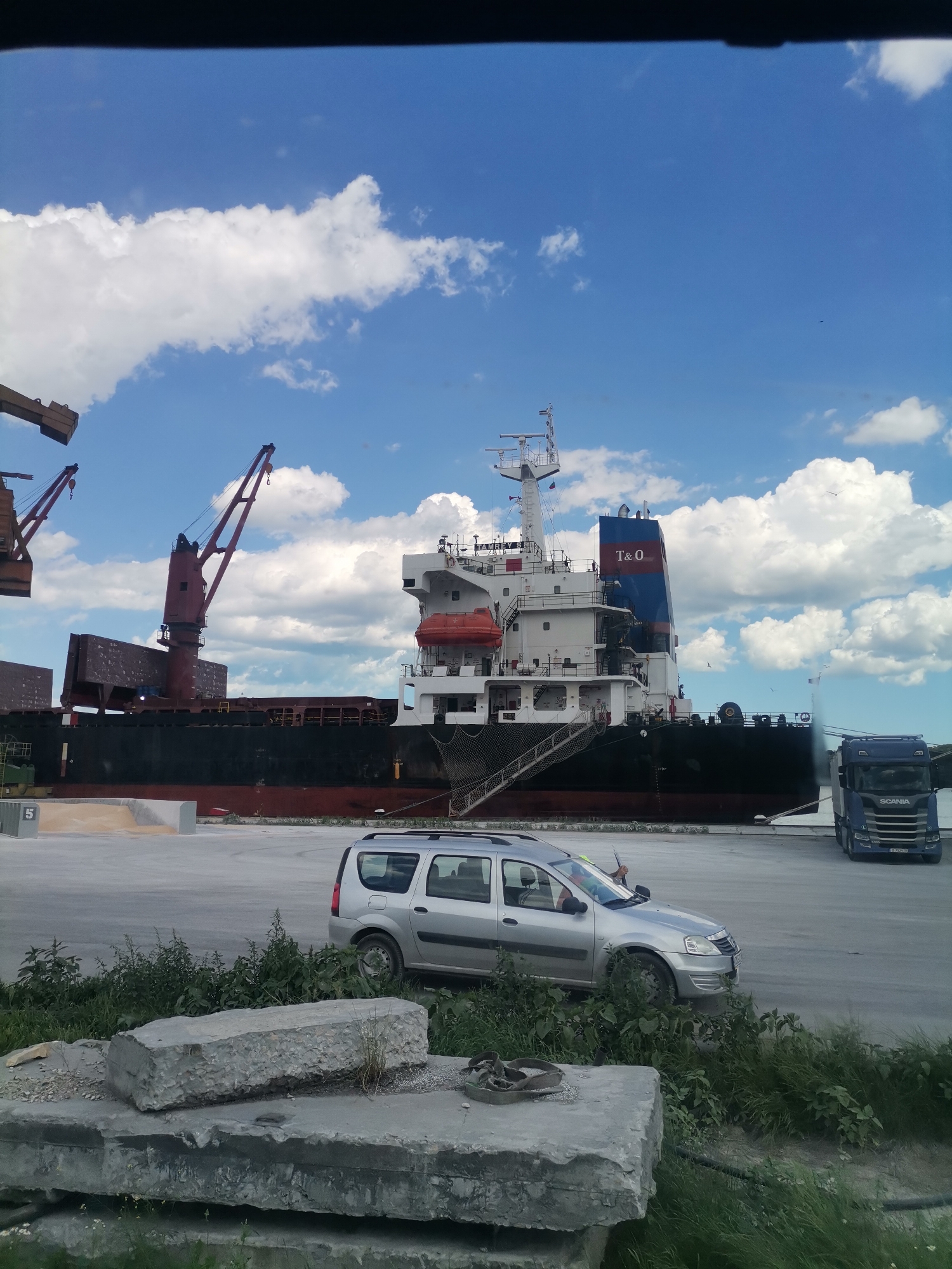 The image depicts a harbor scene in Ezerovo, Bulgaria, under a bright blue sky with scattered white clouds. A large cargo ship is docked, with cranes positioned nearby. In the foreground, a silver minivan is parked on a paved area. There are stacks of concrete blocks and lush green vegetation in the immediate foreground. A blue Scania truck is parked behind the minivan, to the right of the ship. The scene suggests ongoing port activity with the presence of heavy machinery and vehicles.